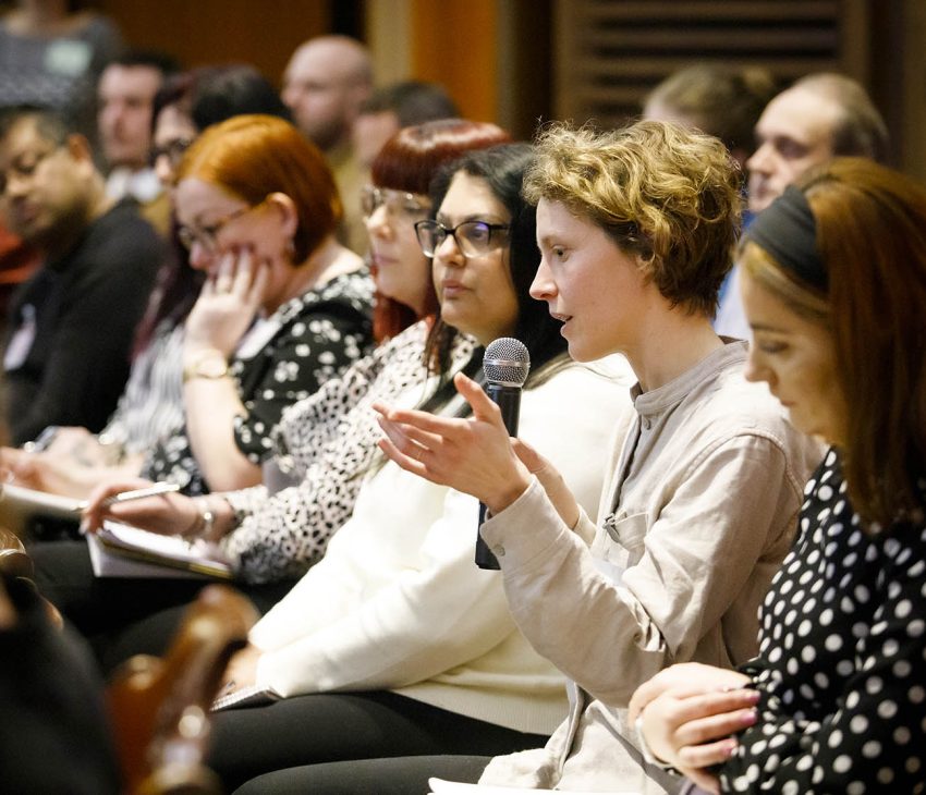 woman holding microphone speaking an event with other people seated besides her