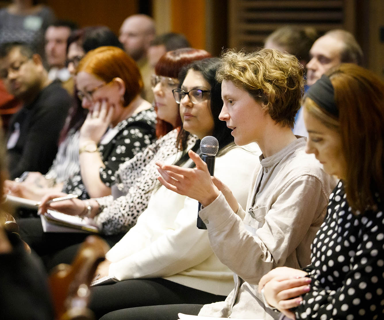 woman holding microphone speaking an event with other people seated besides her