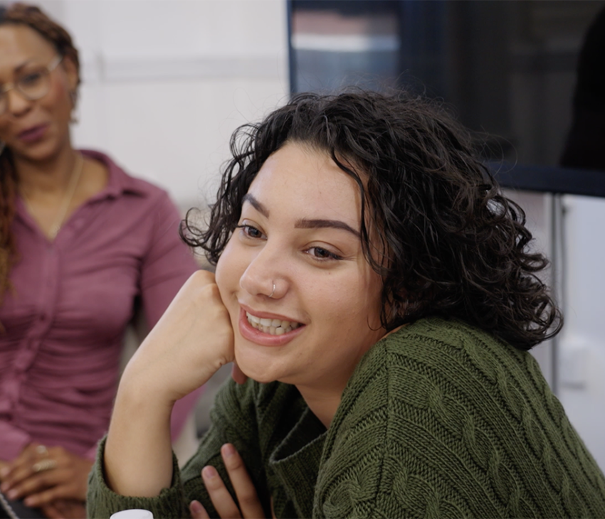 A girl smiling, head resting on her hand, adult looks at her in the background