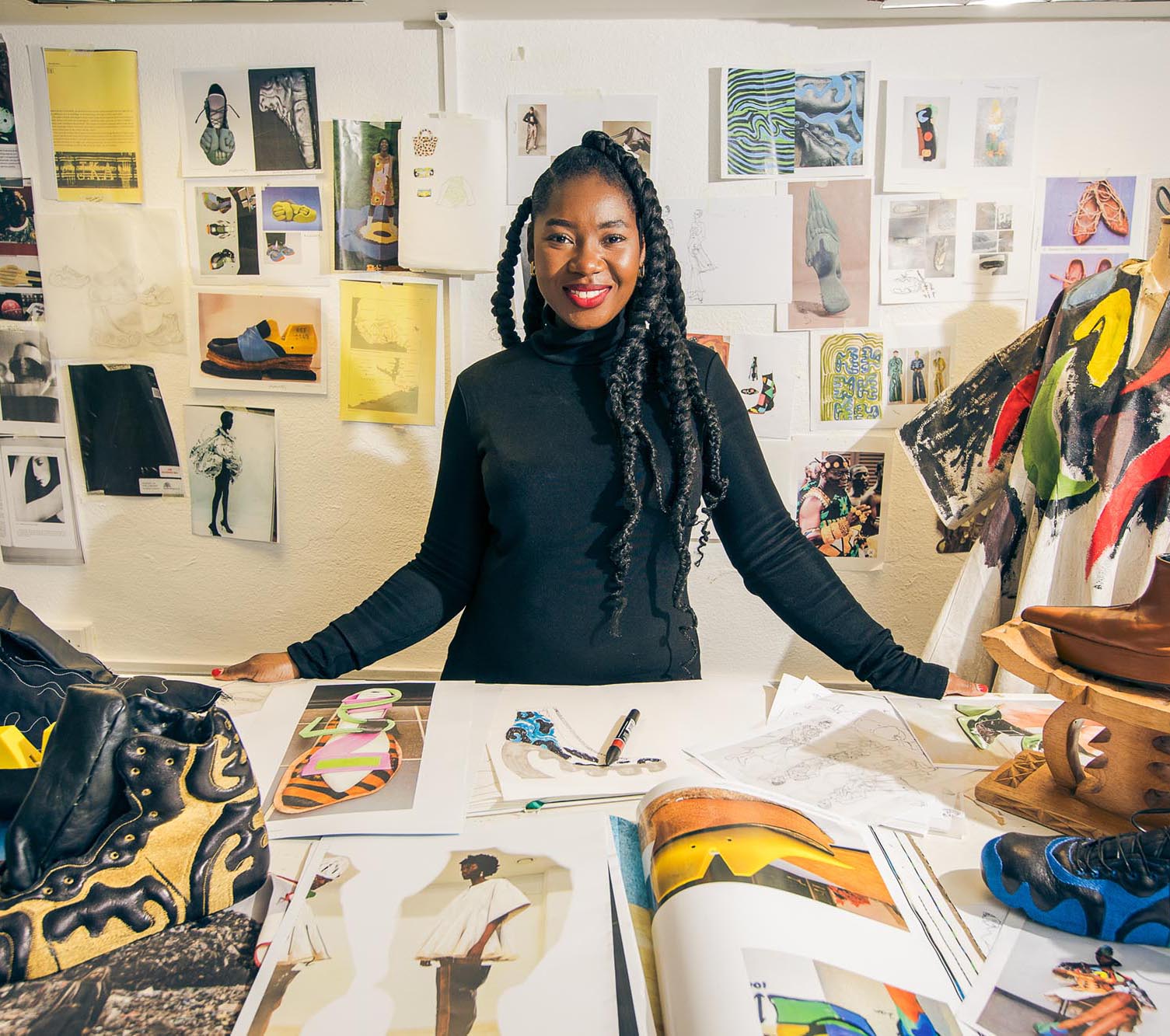 A woman in a black polo neck stands behind a table covered with art supplies, fashion sketches, and design materials, with a wall of photos and inspiration images behind her.