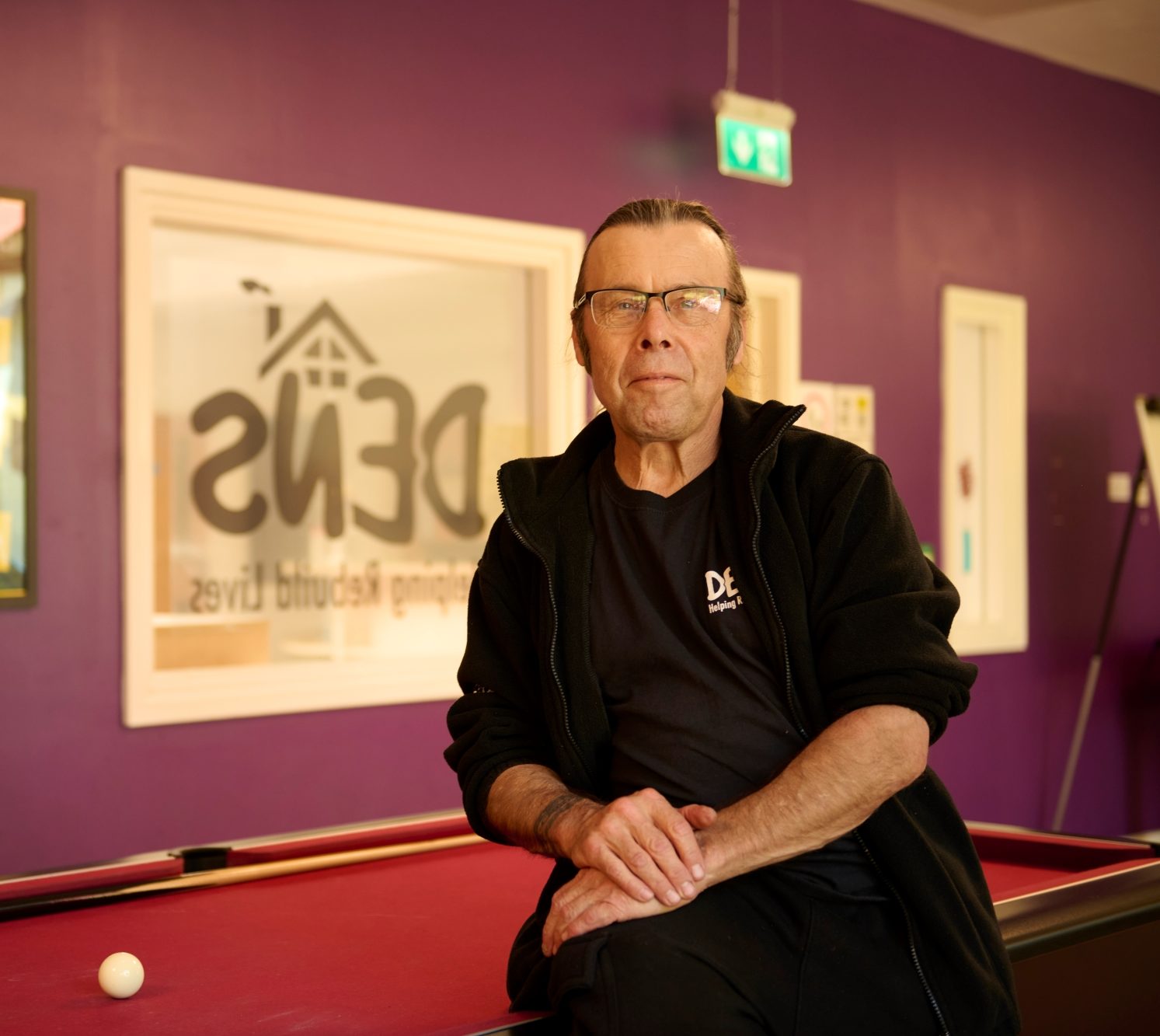 Man sitting on pool table, DENS logo in the background