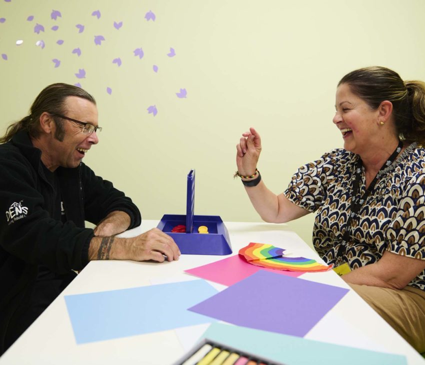 Two adults sit at a table, smiling and playing a tabletop game. Colourful paper sheets and craft supplies are on the table, and purple butterfly decorations are on the light green wall behind them.