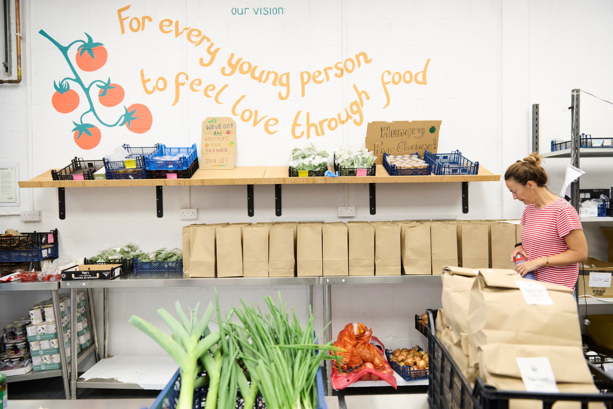 A woman in a striped shirt stands near shelves with brown paper bags and food items. A sign reads, 