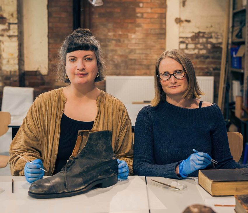 two women stand at a desk with a leather shoe and a leather bound book