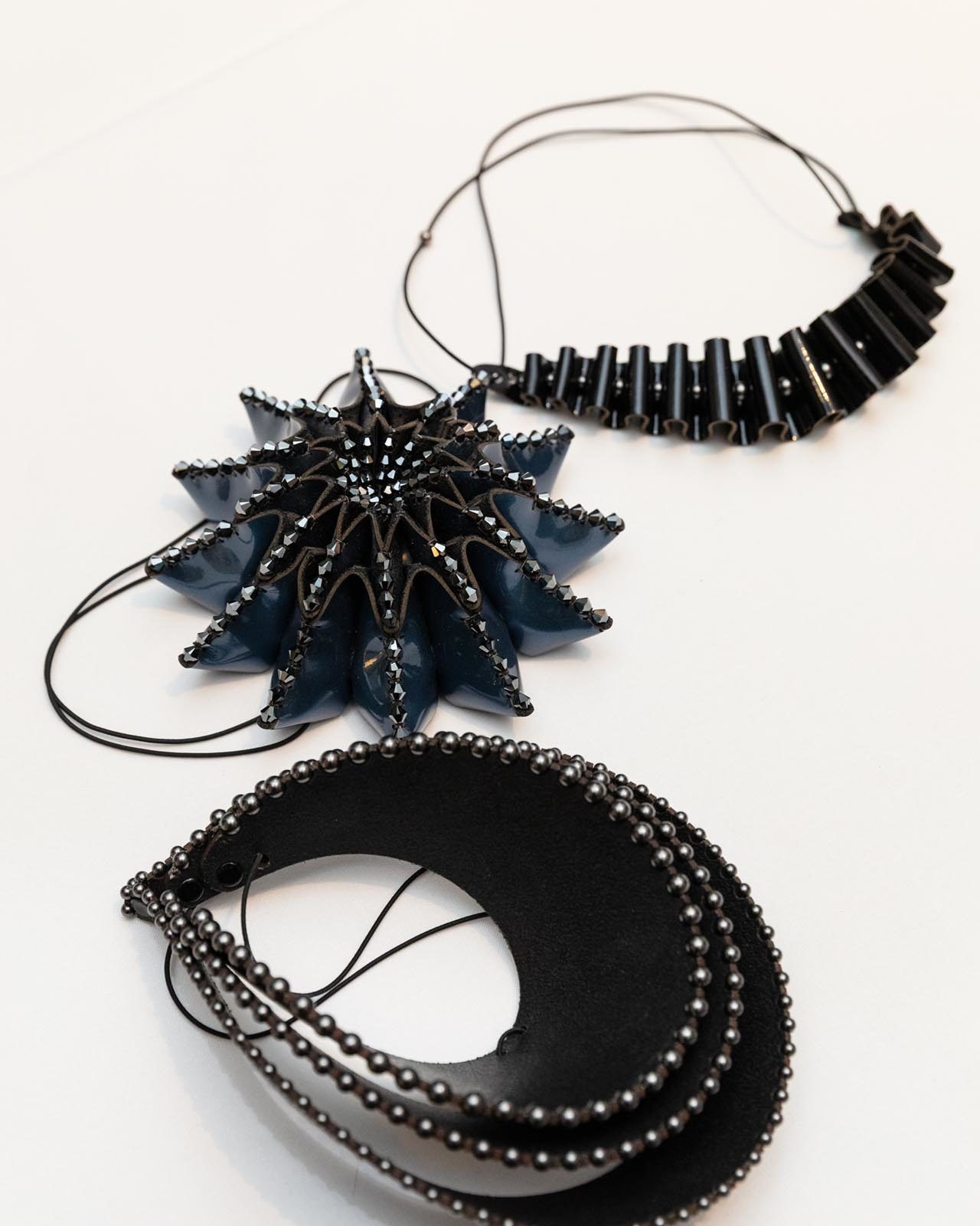 Three black necklaces with geometric designs, inspired by the history of British leather craft, are displayed on a white surface. One has a star-like shape, another features tube-like segments, and the third showcases layered ovals with metallic beads.