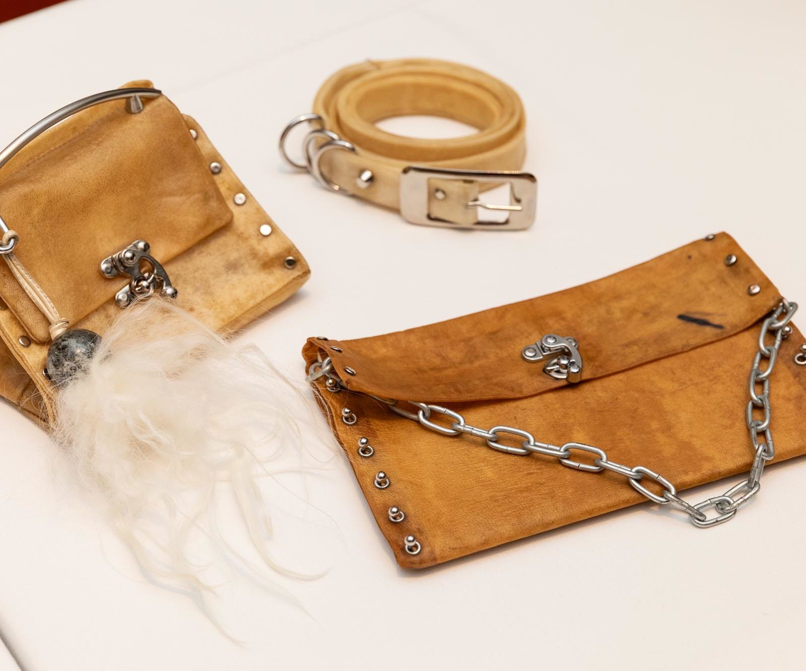 Three tan leather accessories on a white surface; a small handbag with metal studs and furry detail, a matching belt, and a flat pouch with chain and metal clasps.