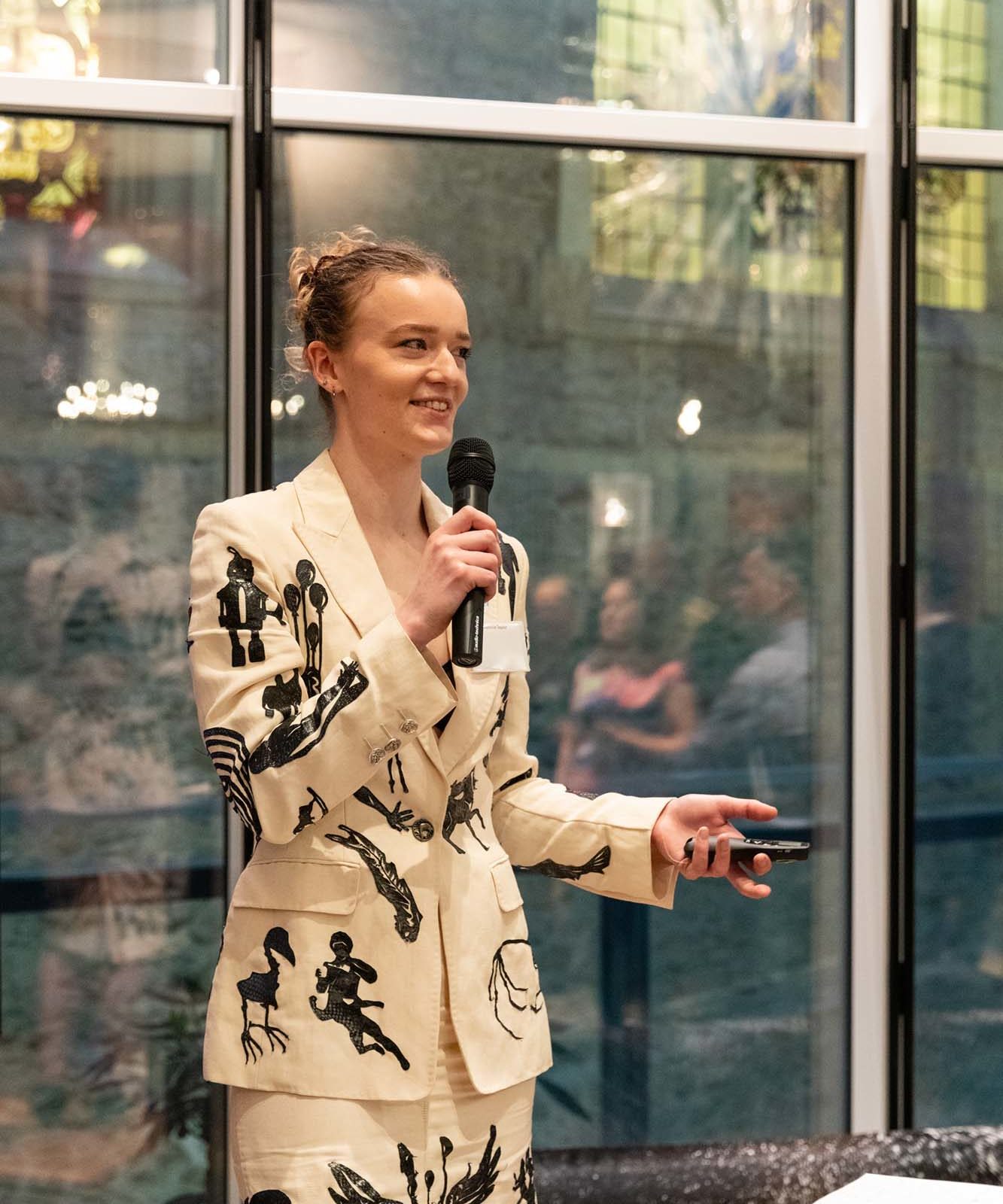 A person wearing a cream-coloured suit with black graphic designs holds a microphone and gestures while speaking indoors, standing in front of large windows with a blurred audience in the background.