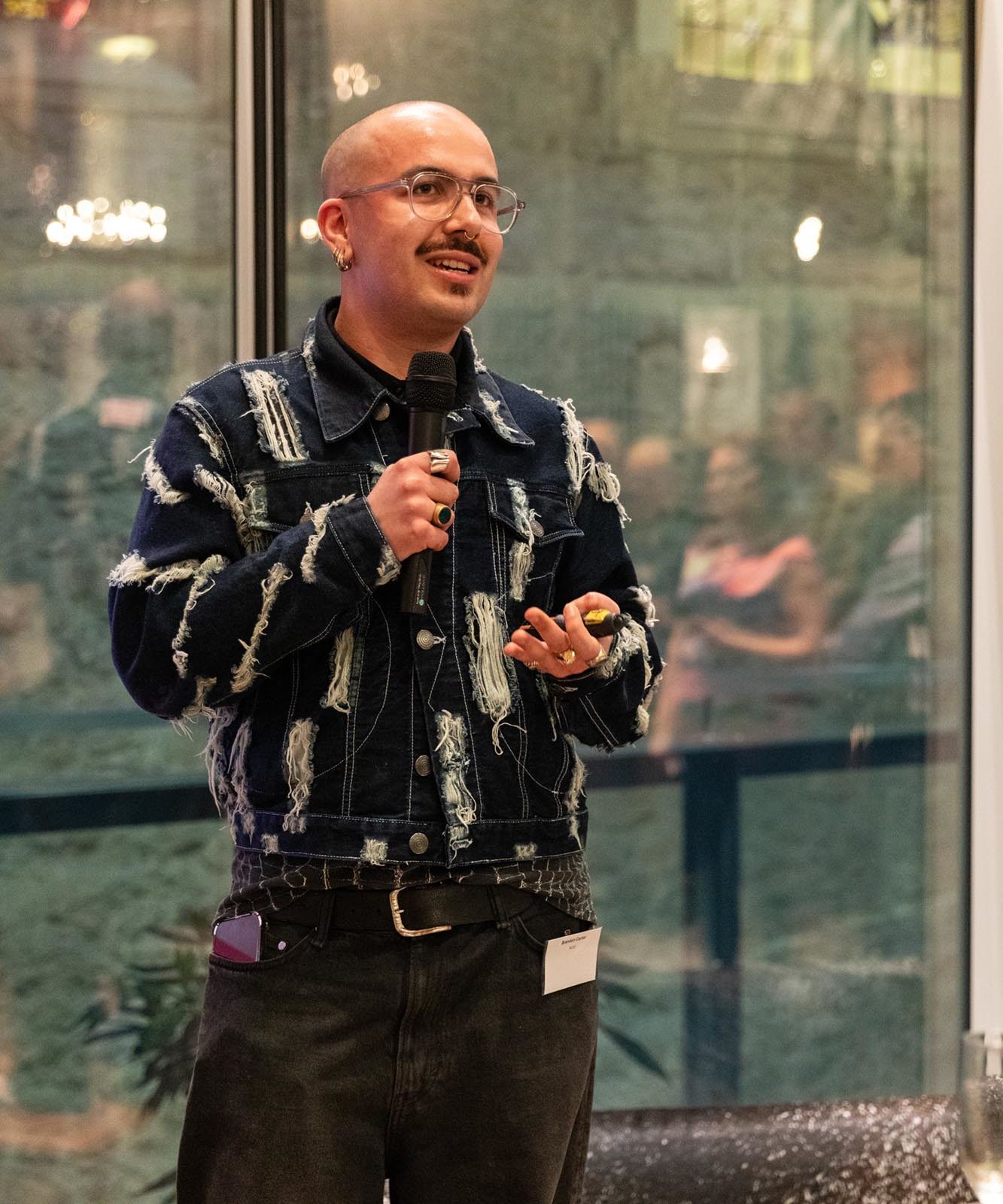 A person with glasses speaks into a microphone indoors. Wearing a ripped denim jacket they stand before an audience.