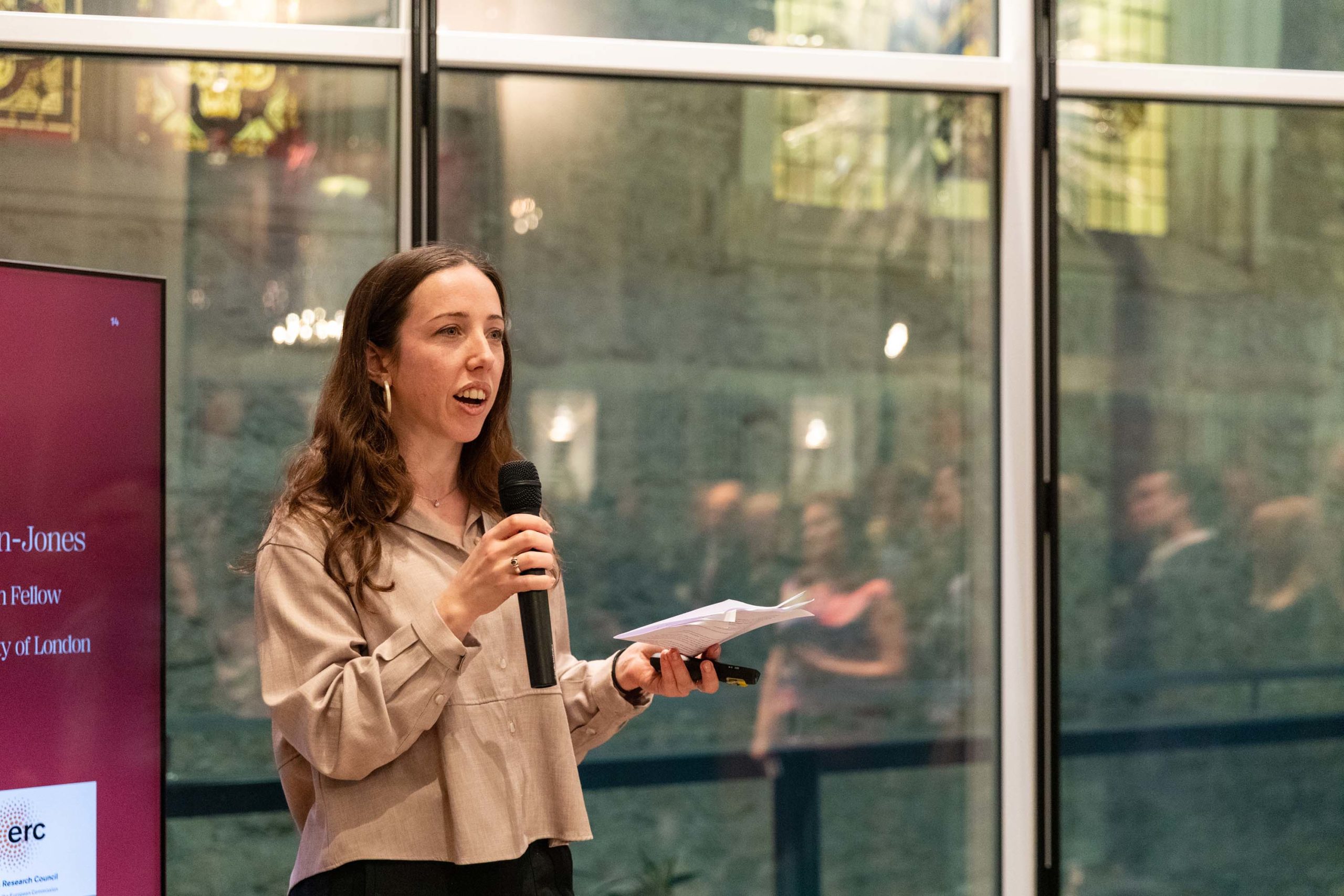 A woman with long brown hair speaks into a microphone whilst holding papers. She stands beside a red screen with text, in front of large glass windows showing blurred people and a stone building outside.