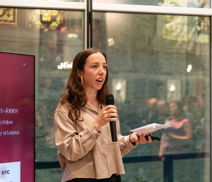 A woman with long brown hair speaks into a microphone whilst holding papers, presenting. She stands indoors near large windows, with blurred people and lights in the background. A red presentation screen is on her left.