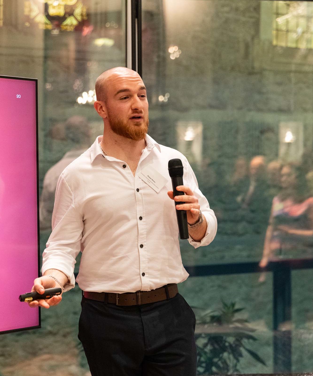 A man in a white shirt and dark trousers holds a microphone and a presentation remote, standing in front of a screen, speaking to an audience in a modern indoor setting.