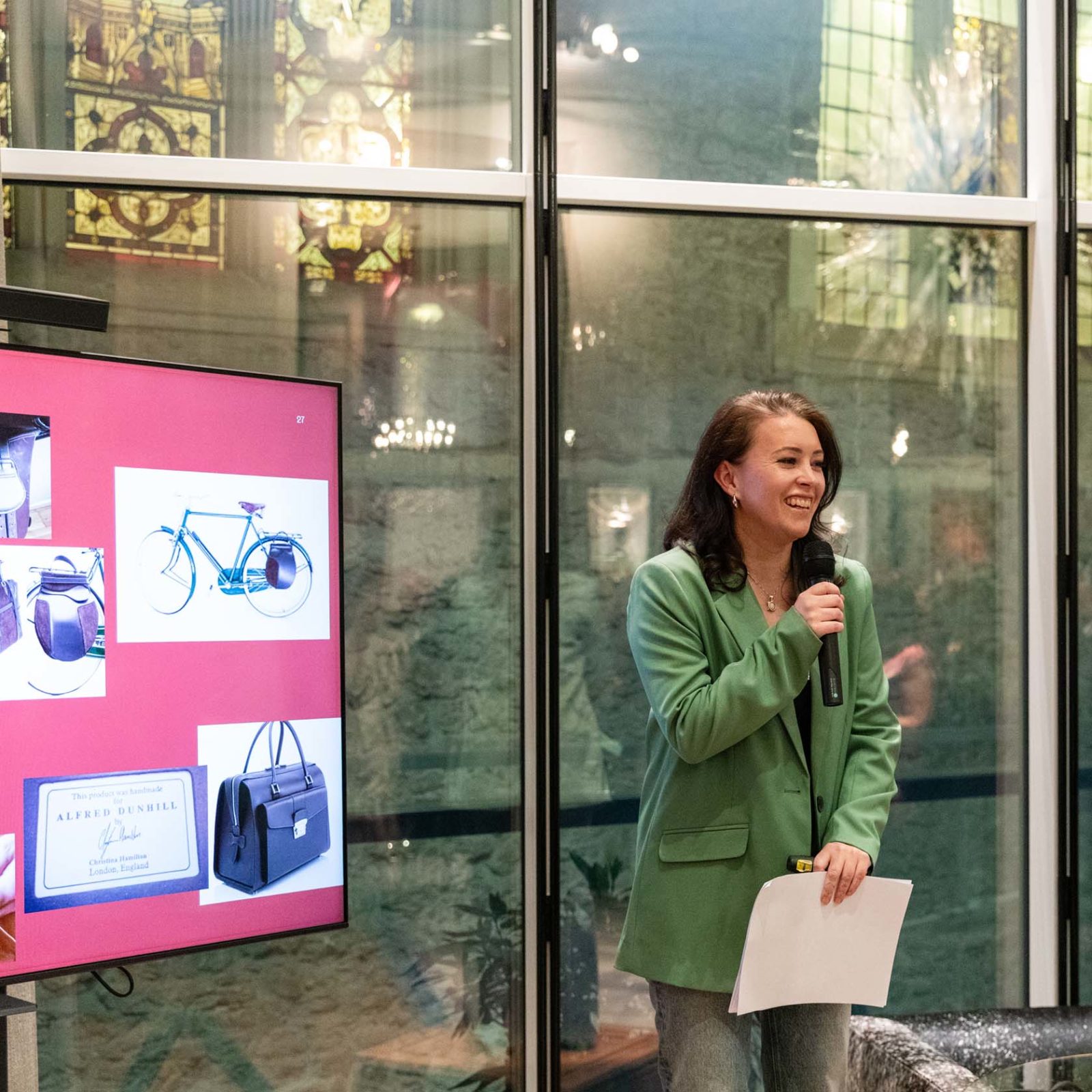 A person stands indoors holding a microphone and papers, speaking in front of a screen displaying images of bags, a bicycle, and a business card. Stained glass windows and reflections are visible in the background.