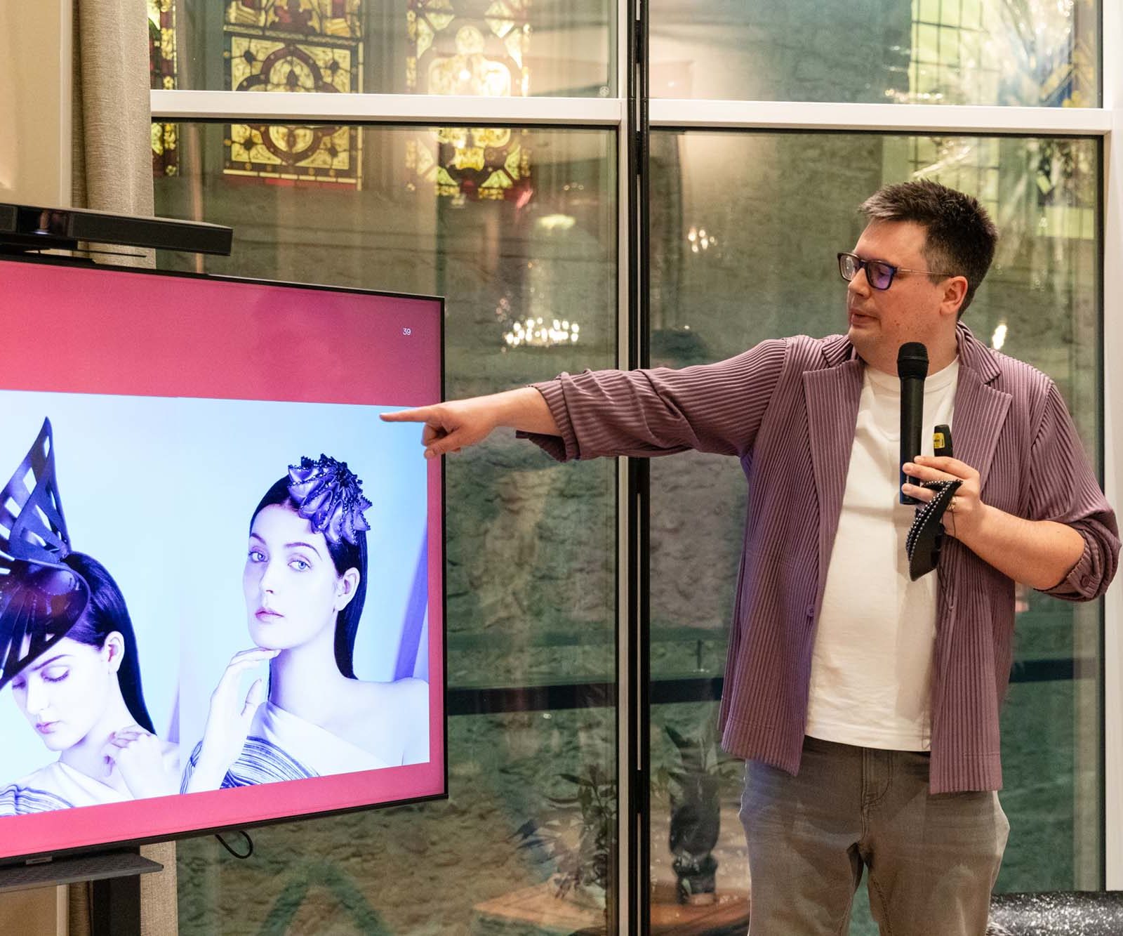 A man holding a microphone points at a screen displaying two women wearing geometric leather headpieces. He stands indoors in front of large windows with decorative glass panes.