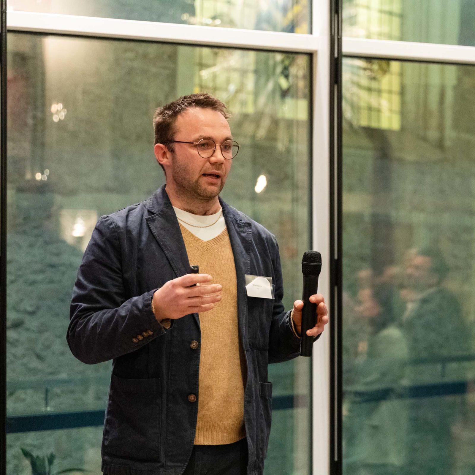A man wearing glasses, a blue jacket, and a tan jumper holds a microphone whilst speaking indoors in front of large windows.