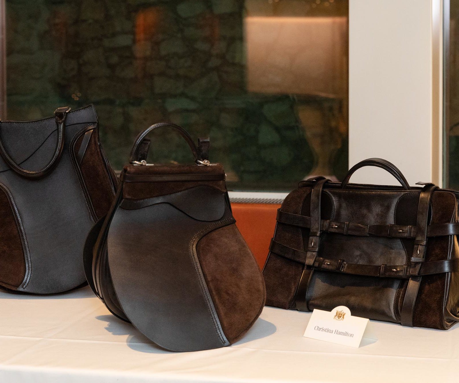 Three dark brown leather bags are displayed on a white tablecloth. Two resemble saddlebags, and one is a rectangular duffle bag.