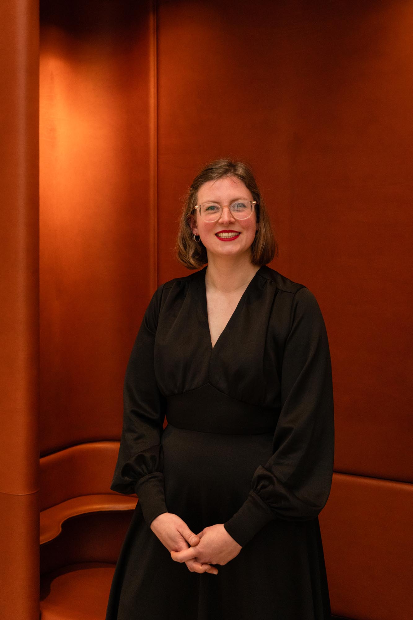 A woman with shoulder-length brown hair, wearing glasses and a black dress, stands smiling in front of a brown leather curved wall and seating area.