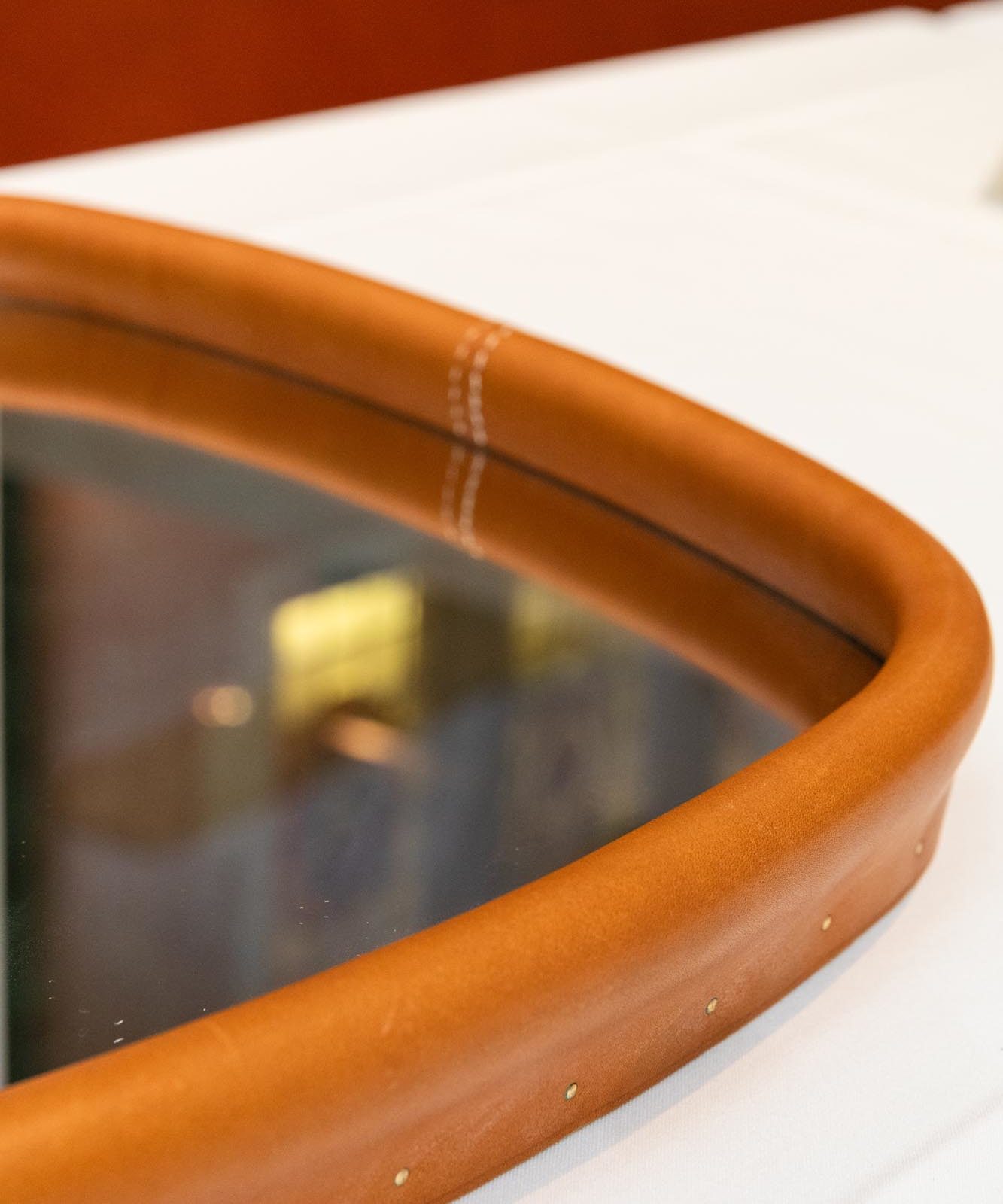 Close-up of a mirror with a rounded, brown leather frame featuring visible stitching and metal rivets.