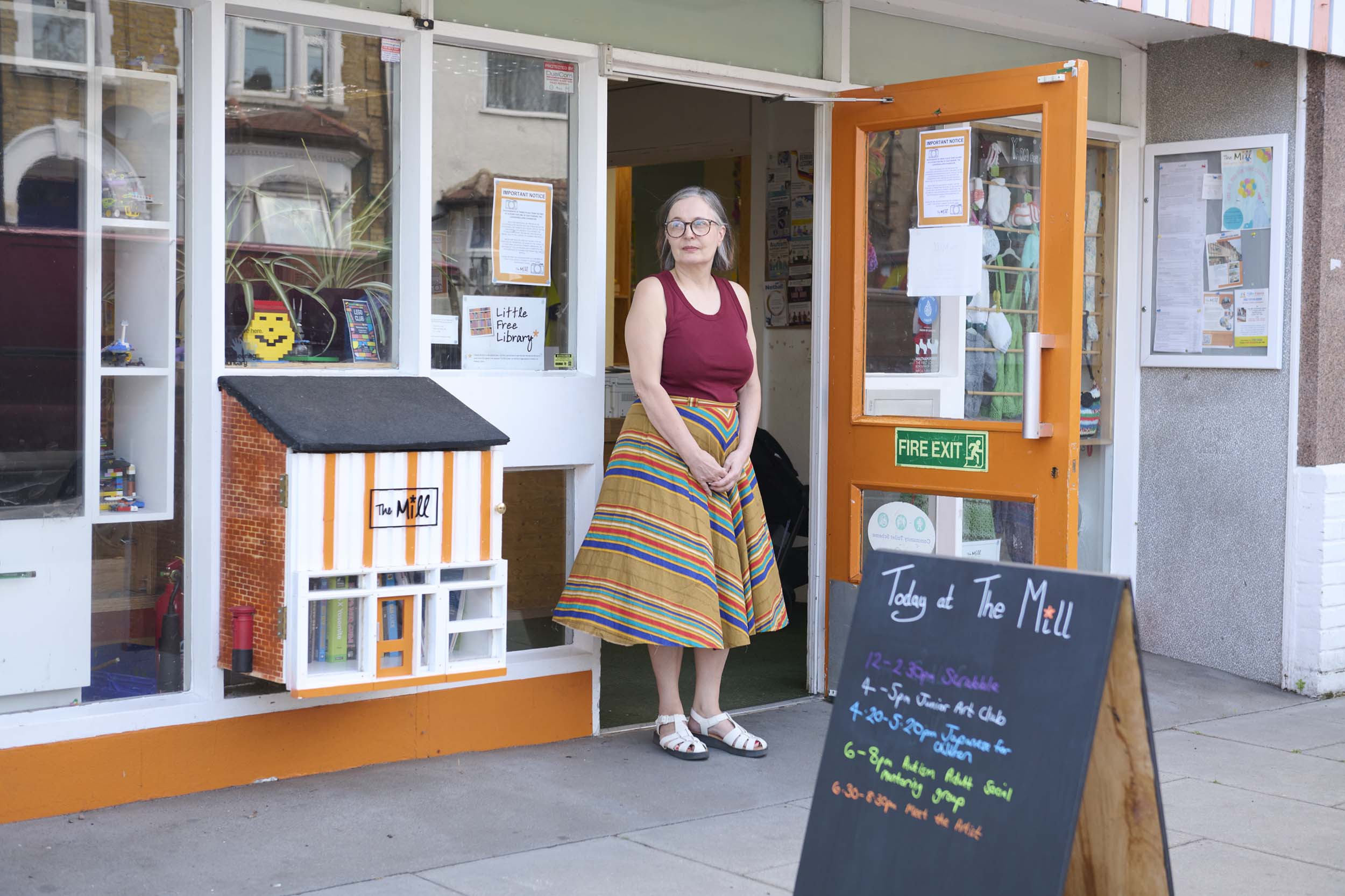A woman wearing glasses, a red sleeveless top, and a colourful striped skirt stands in the entrance of a shop with a chalkboard sign listing events at The Mill and a decorative house display by the door.