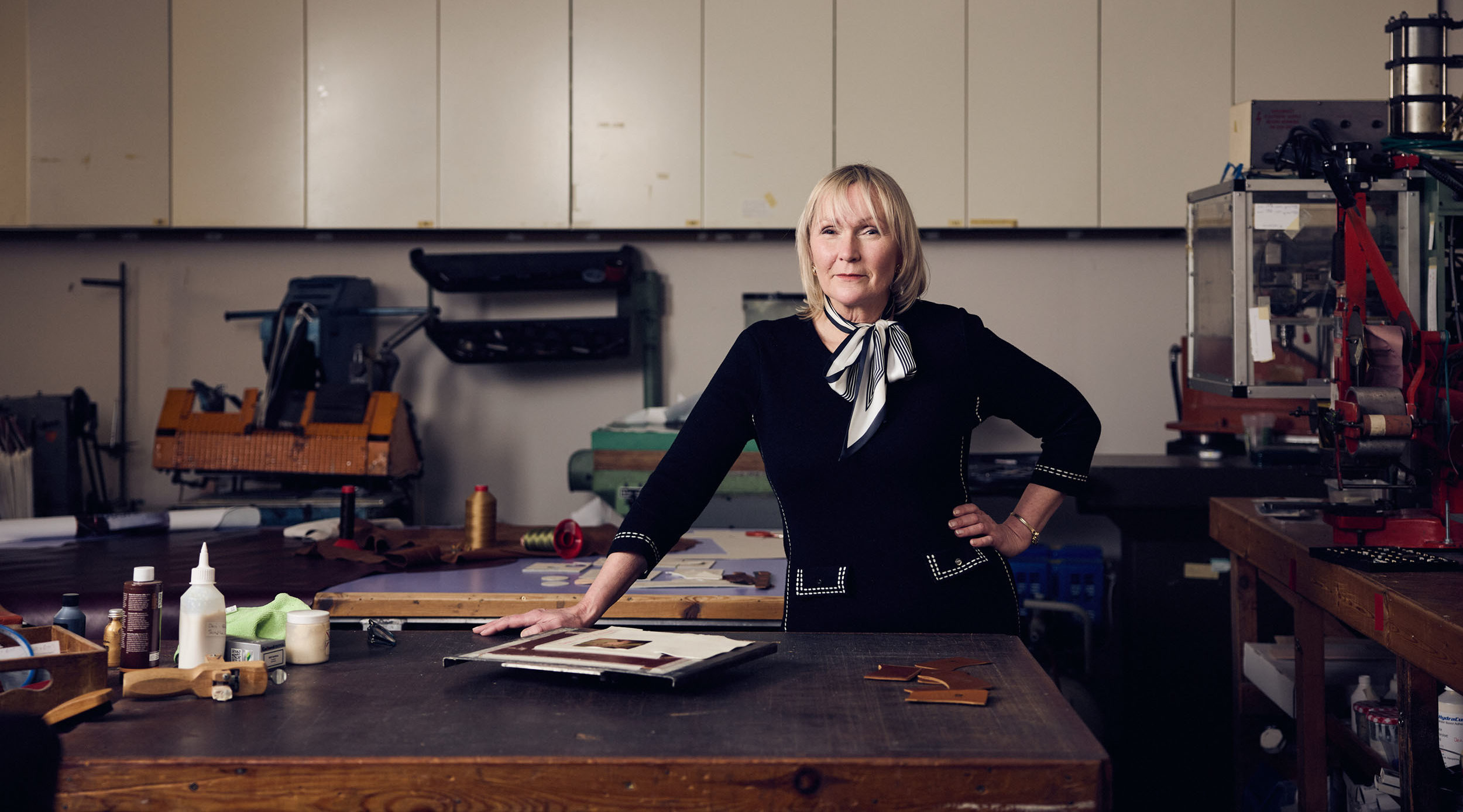 A woman stands with one hand on a workbench in a workshop. The bench is covered with tools, bottles, and materials. Behind her are shelves, cupboards, and various equipment against the wall.