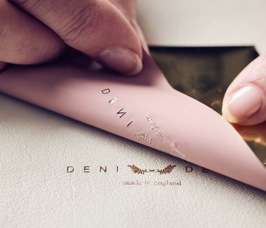 A close-up of two hands peeling back a pink sheet with gold foil, revealing embossed gold text on a beige leather surface. The text partially reads DENI and made in England.