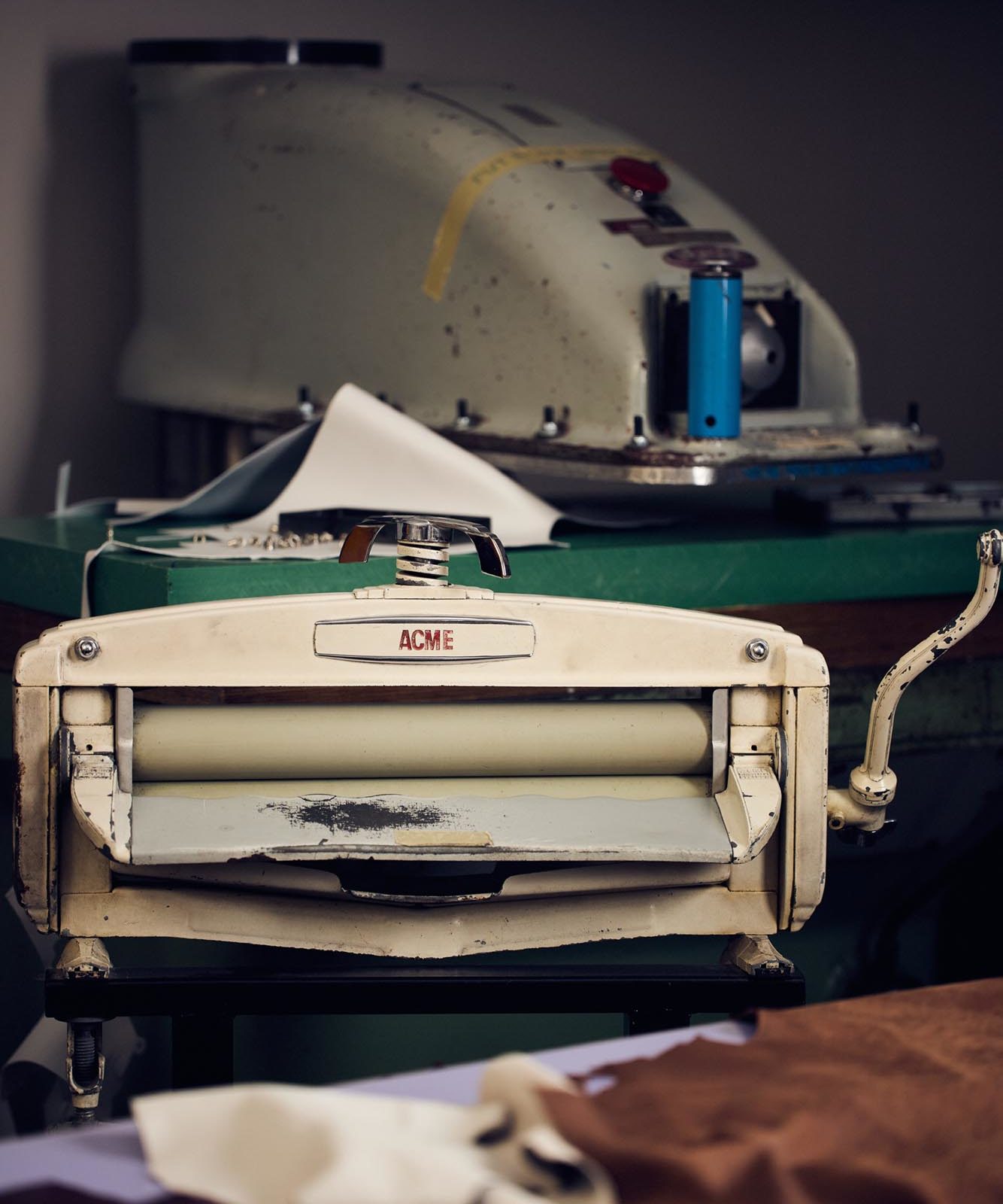 A vintage ACME brand ironing or pressing machine sits on a table with various pieces of fabric and paper around it. Another large machine and tools are visible in the background.