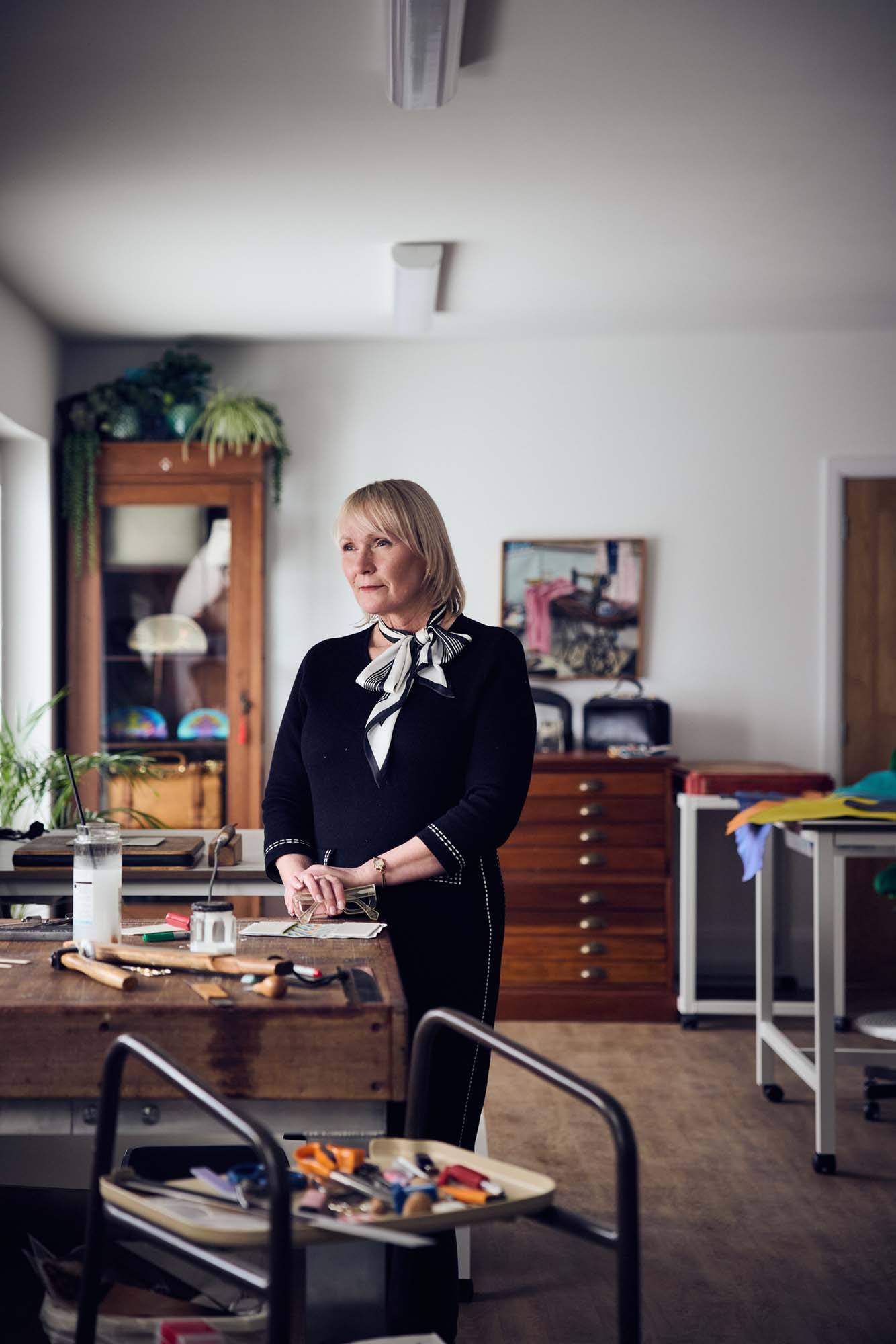 A woman stands in a well-lit workshop, wearing a black dress with a white scarf. She is near a workbench with tools and materials scattered on it. Cupboards, plants, and artwork are visible in the background.