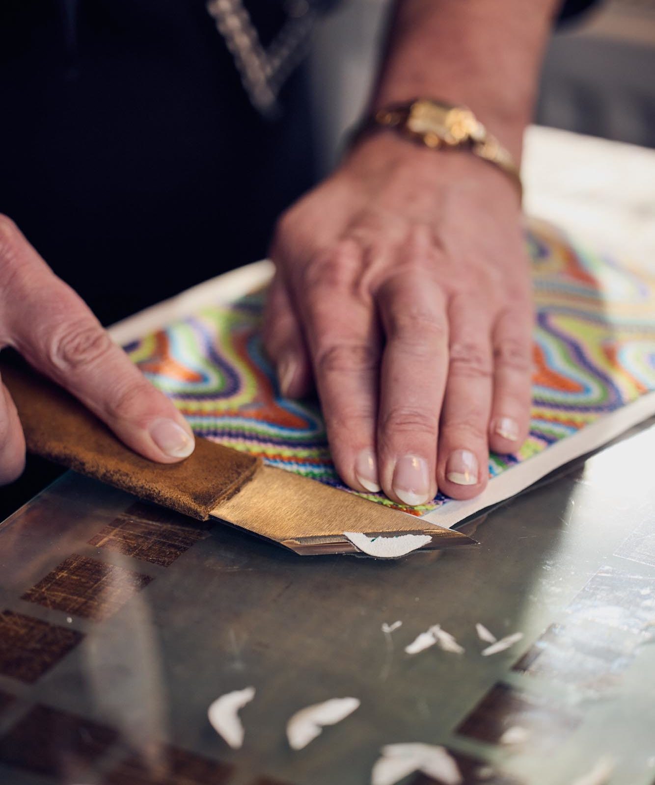 A person uses a tool to cut a piece of material with a colourful, wavy pattern on a glass surface. The person's hands are visible, showing a bracelet and neatly manicured nails.