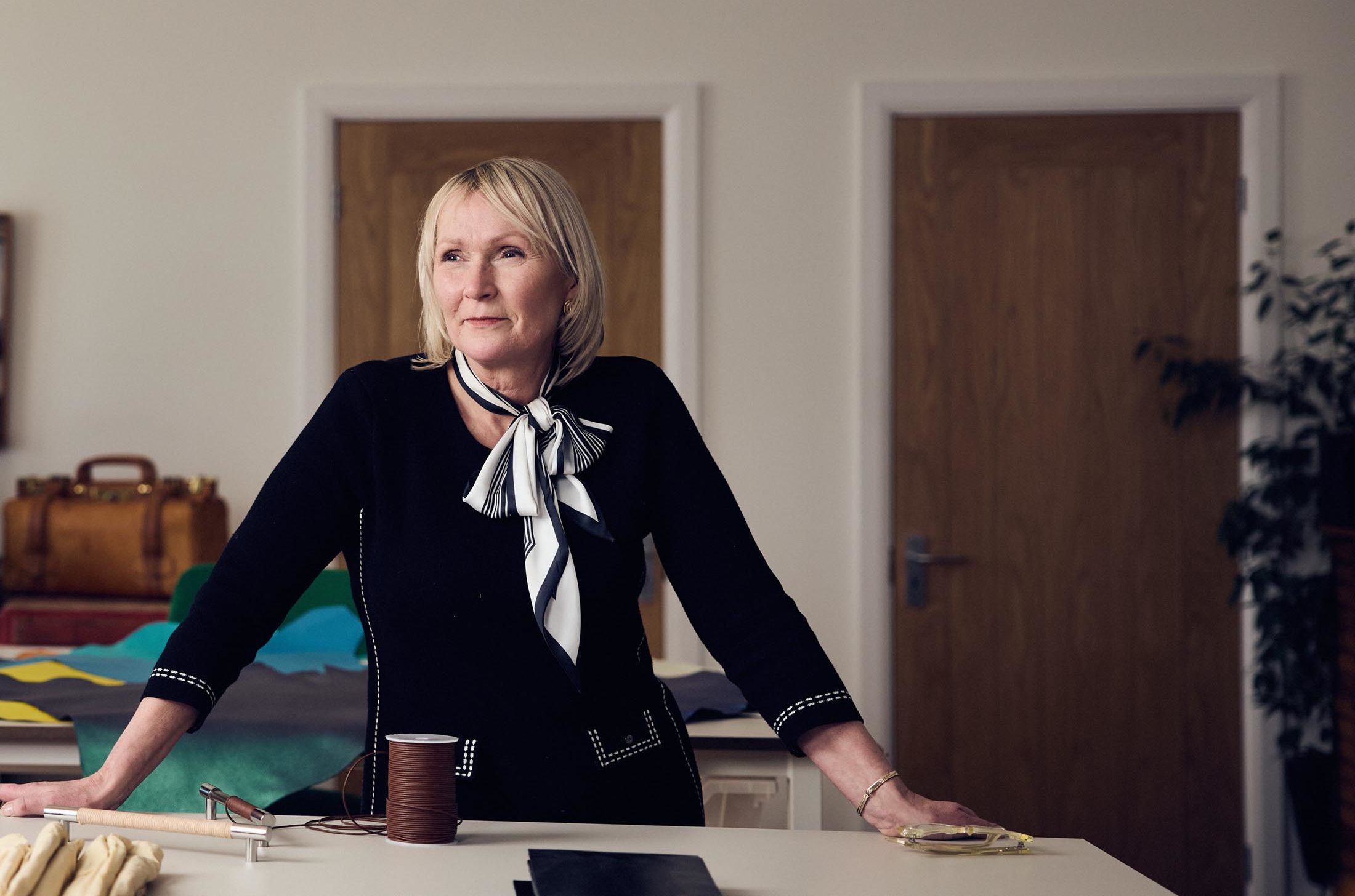 A woman with blonde hair wearing a black outfit and a patterned scarf stands at a table in a well-lit room with wooden doors, some bags, and sewing materials visible in the background.