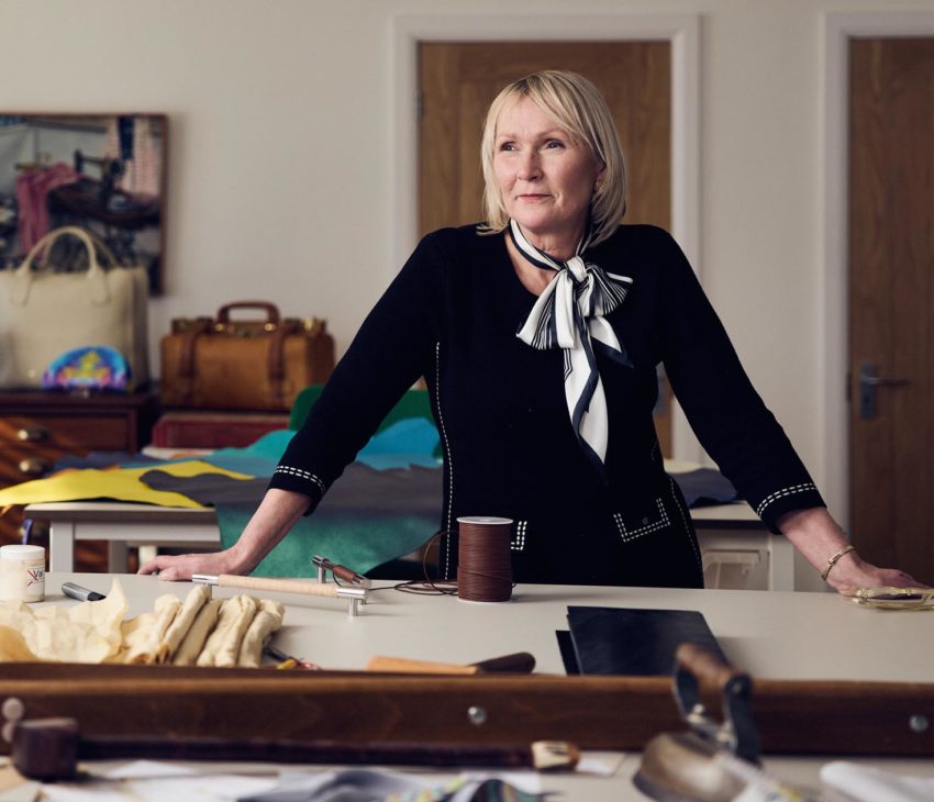 A woman stands behind a worktable covered with tools and materials in a workshop or studio space. She wears a black top with a scarf and looks thoughtfully to the side. Shelves, bags, and a colourful globe are in the background.
