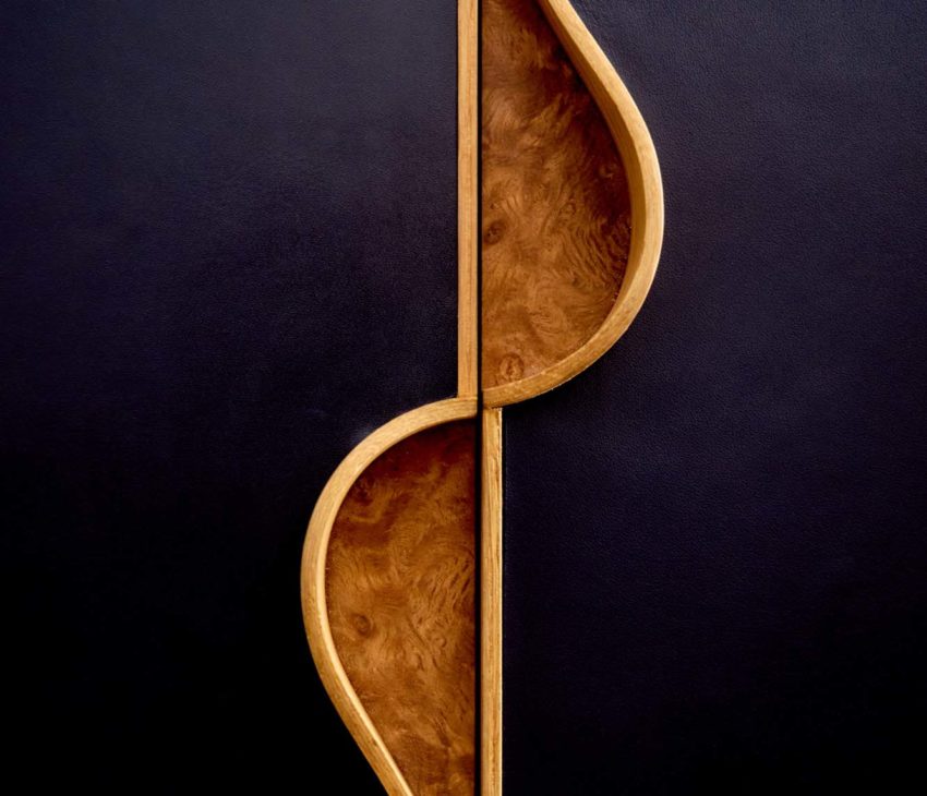 A close-up view of two wooden handles with curved, wave-like edges, set against a black leather background. The wood has a natural finish, showing its grain and texture. The doors meet at the centre, creating a symmetrical design.