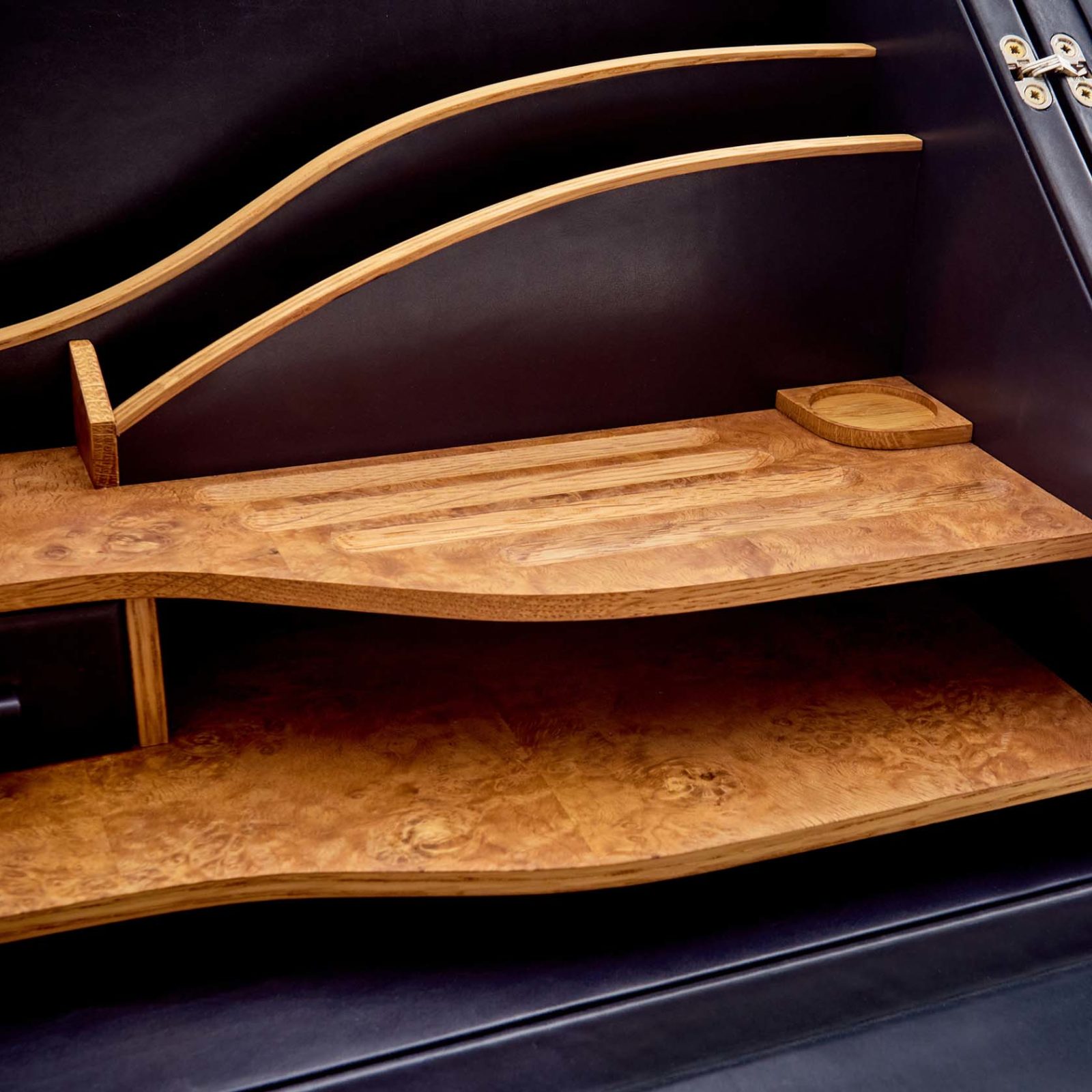 Two wooden shelves with a wavy design are installed against a dark wall. The upper shelf has curved wooden supports and a rectangular inset, whilst the lower shelf has a natural wood grain pattern.