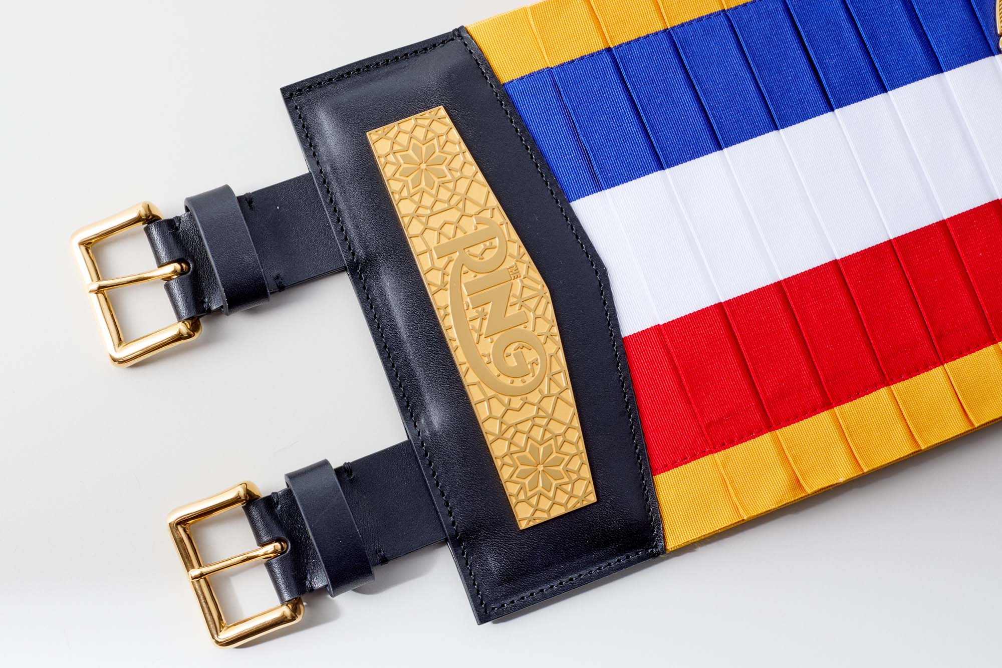 A close-up of a colourful pleated fabric belt with horizontal blue, white, red, and yellow stripes. The belt has gold-tone buckles and a black leather section with a gold embossed geometric pattern and stylised emblem.