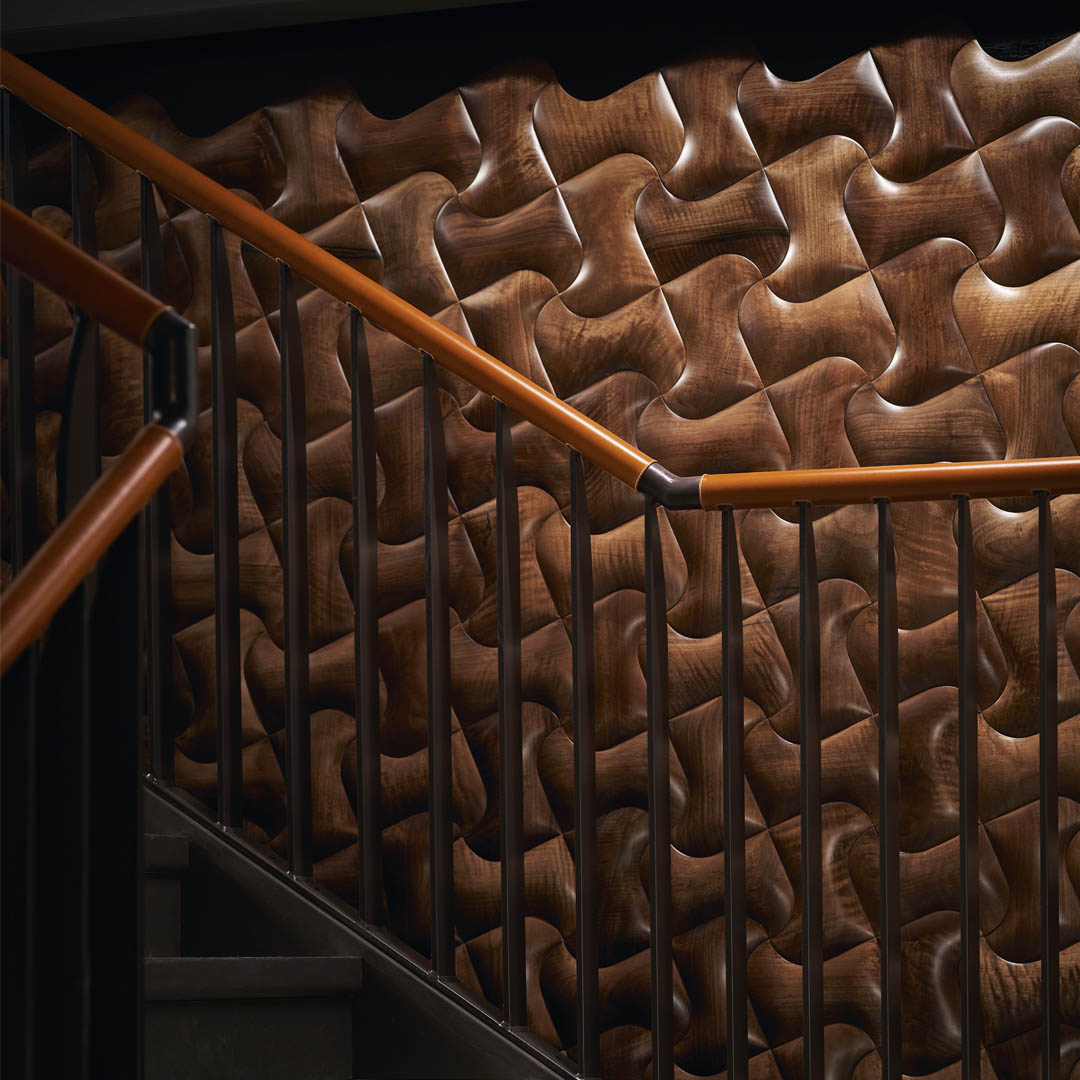 A staircase with leather railings and wooden handrails is set against a textured, dark wood wall featuring an interlocking, puzzle-like pattern. The lighting accentuates the wall’s three-dimensional, glossy surface.