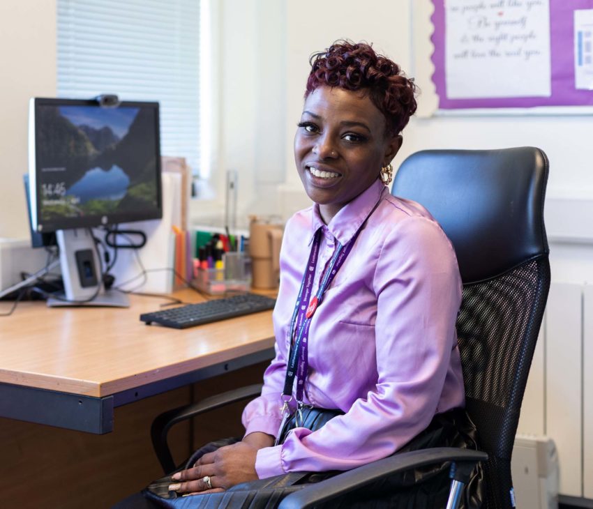 A woman wearing a light purple blouse and lanyard sits smiling in an office chair at a desk with a computer, pens, and papers in a brightly lit office.