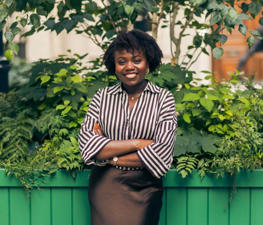 A woman stands in front of lush green plants, smiling with arms crossed. She wears a black and white striped shirt and a dark skirt. There is a green planter box and leafy shrubs in the background.
