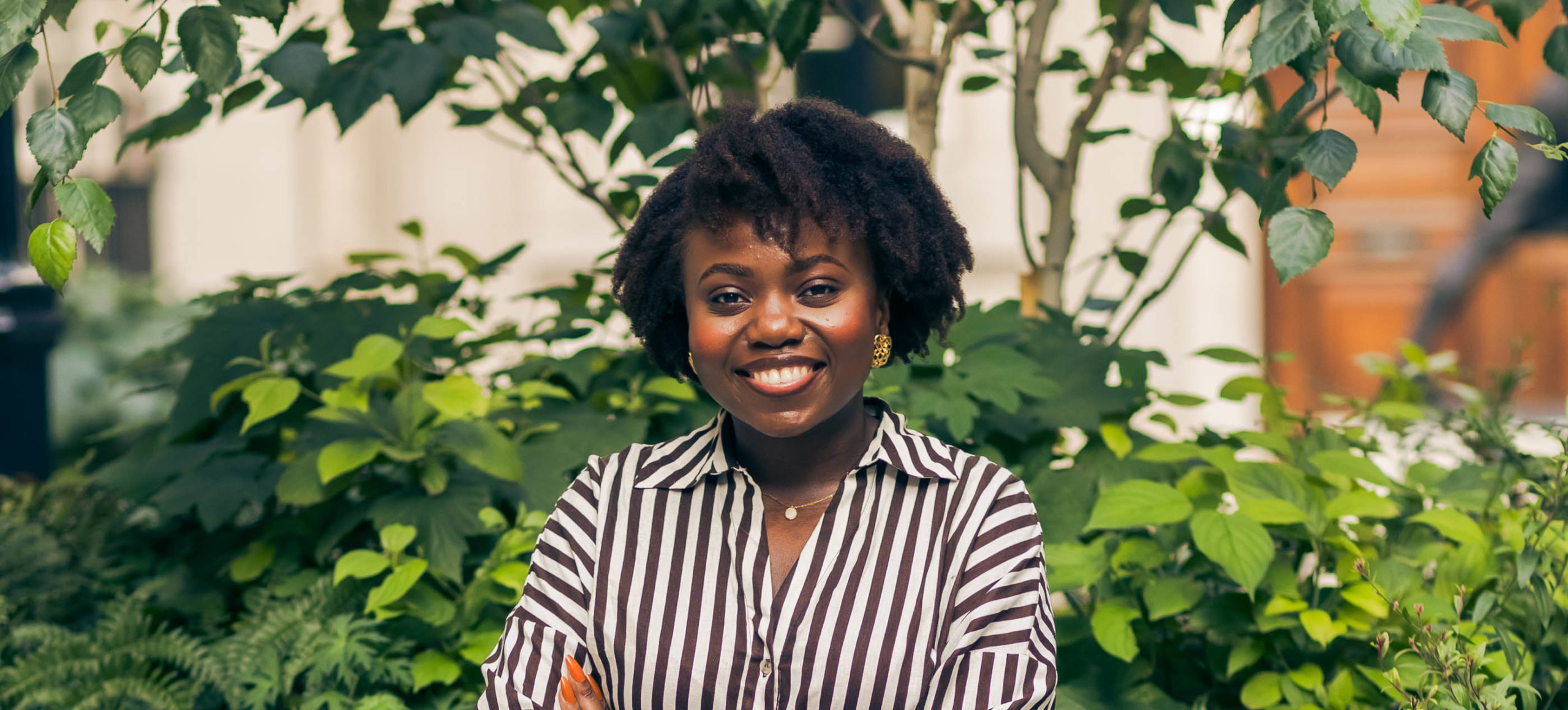 A woman smiles at the camera whilst standing in front of green plants. She is wearing a black and white vertically striped shirt and has her arms folded.