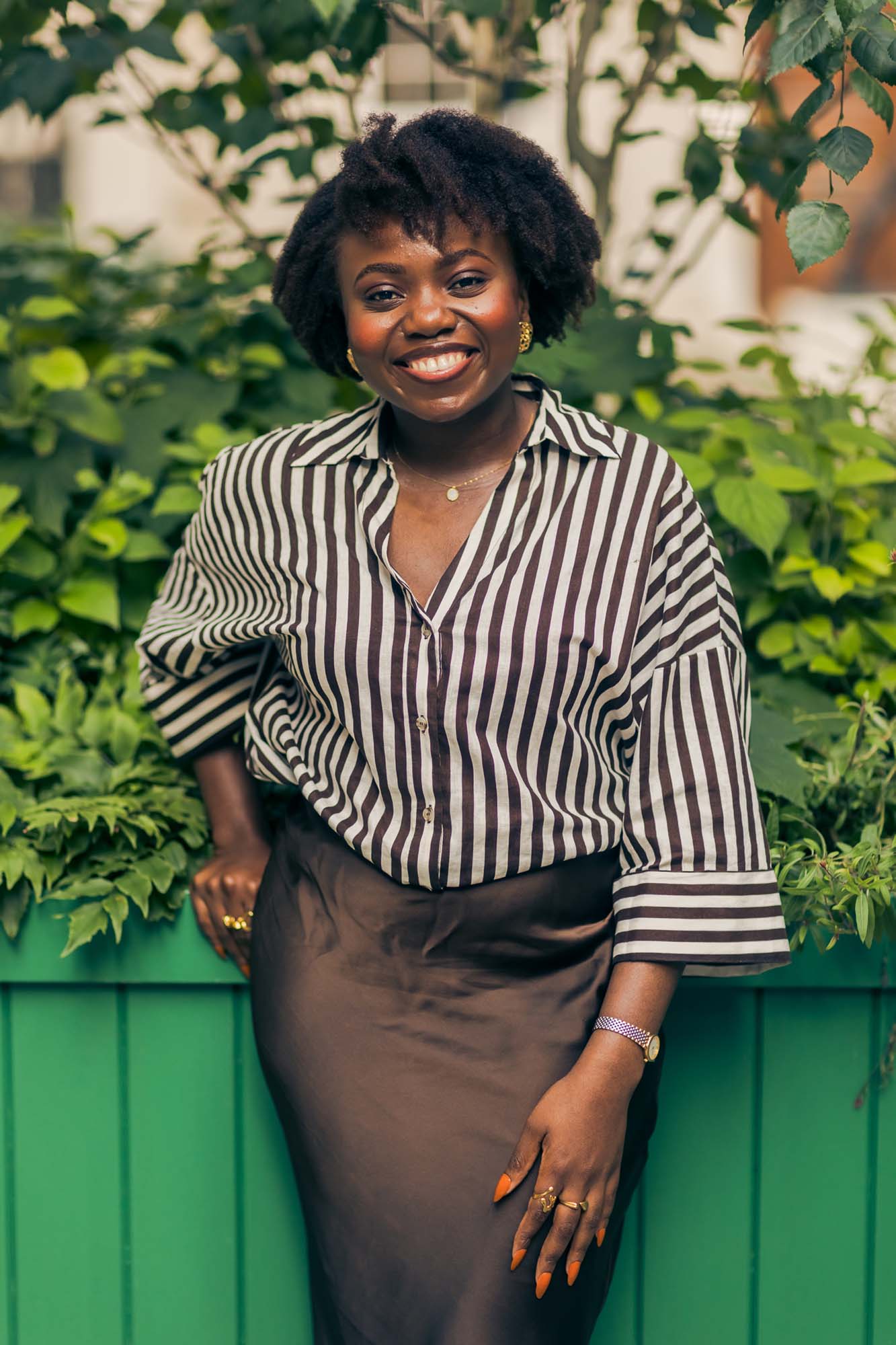 A woman stands in front of lush green plants, smiling with arms crossed. She wears a black and white striped shirt and a dark skirt. There is a green planter box and leafy shrubs in the background.