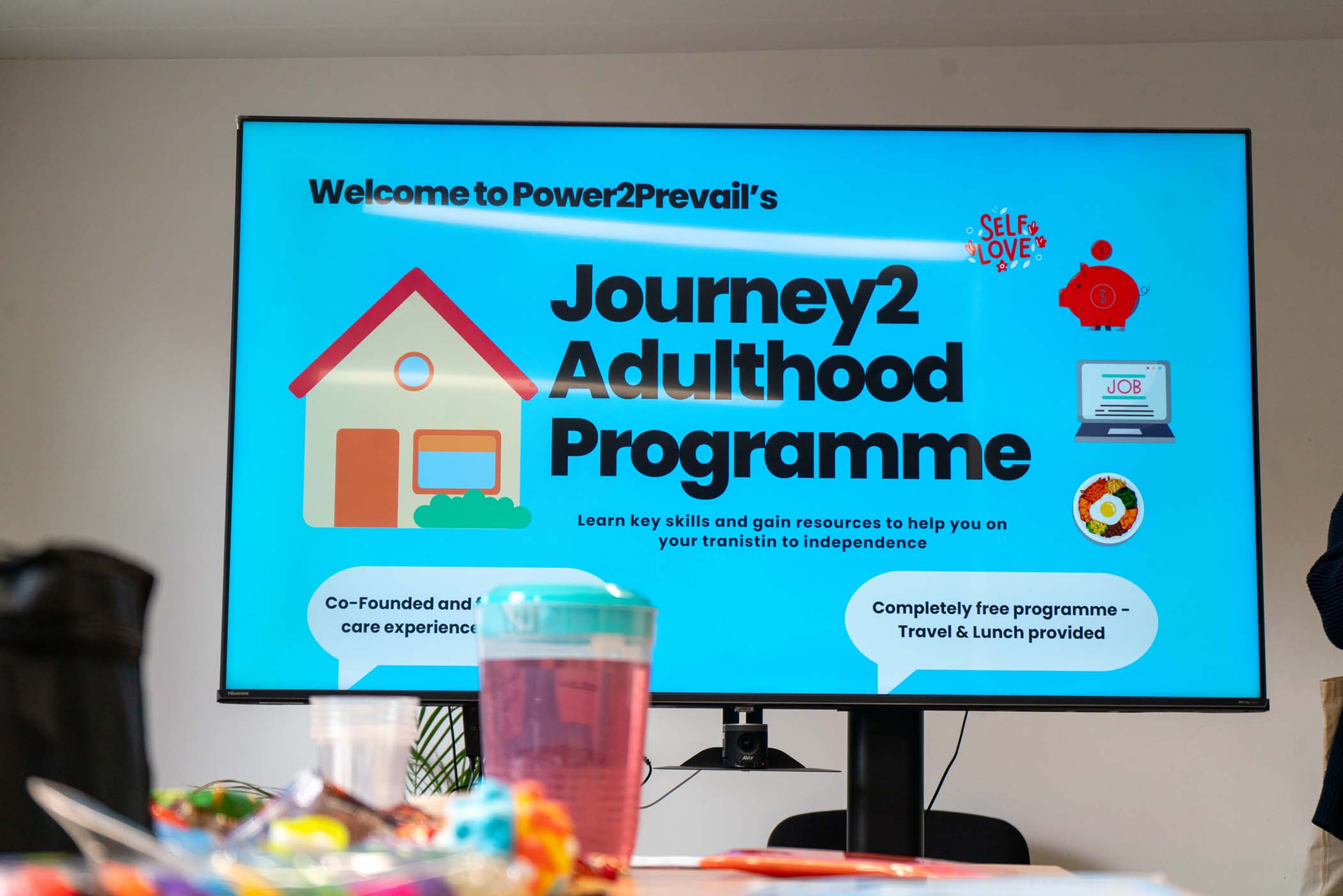 A large screen displays a presentation slide titled Journey2 Adulthood Programme with icons and text about learning key skills for independence. Items and snacks are visible on a table in the foreground.