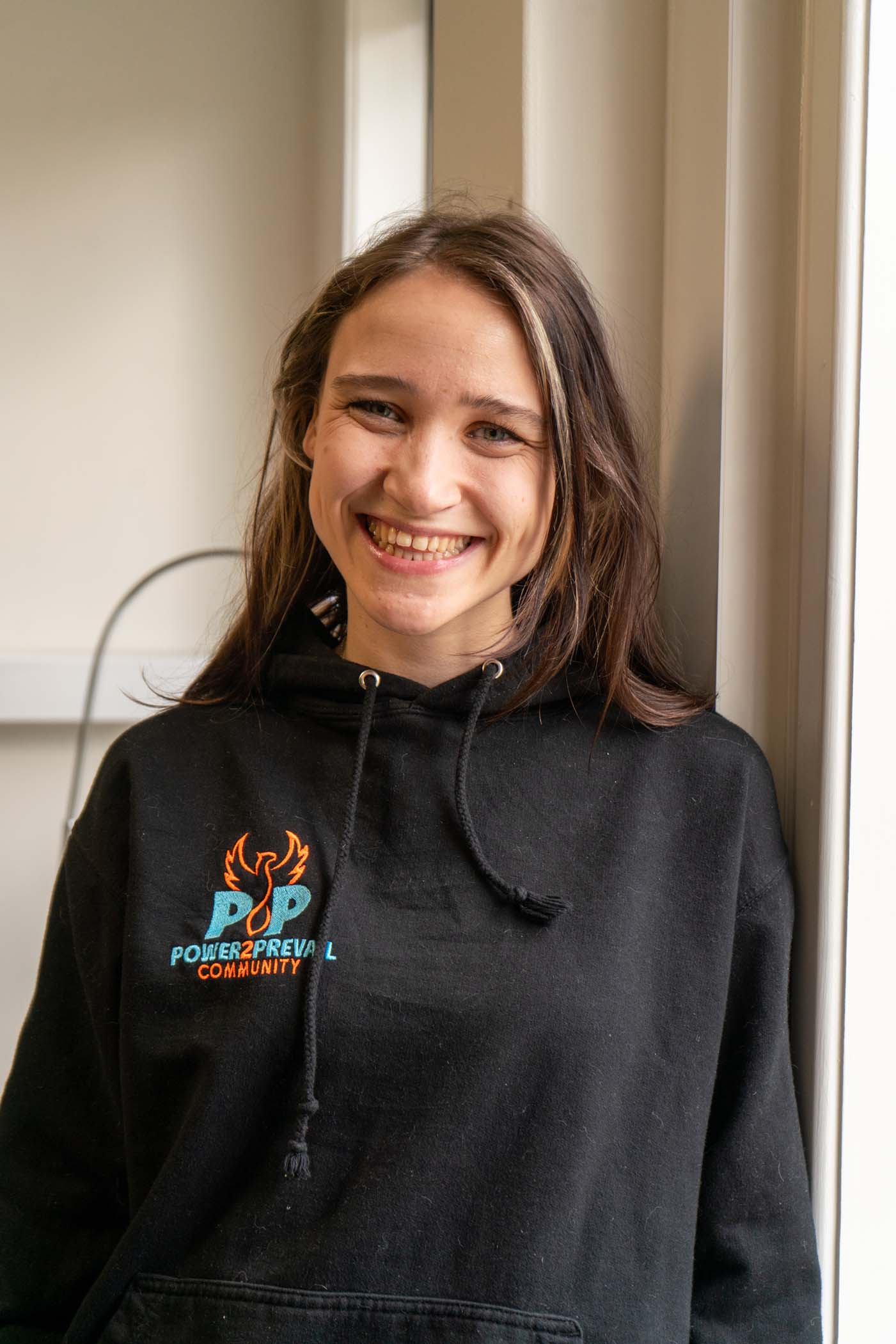 A person with long brown hair smiles whilst standing indoors by a window. They wear a black hoodie with the words POWER 2 PREVAIL COMMUNITY and a graphic of flames on the front.