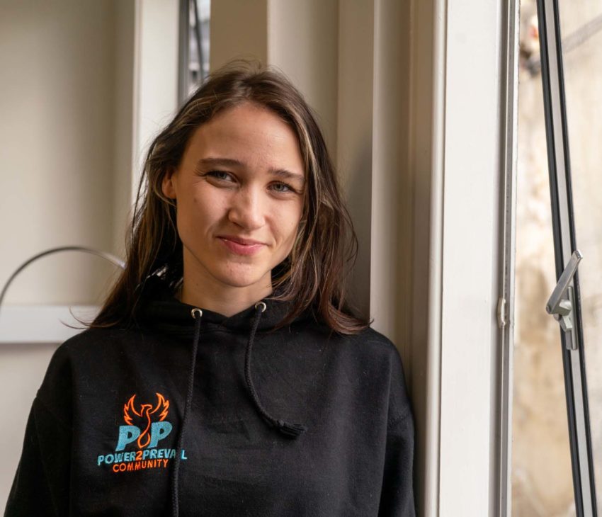 A woman with long brown hair wearing a black “Power2Prevail Community” hoodie stands indoors by a window, looking at the camera with a slight smile. The window is open, and part of a building is visible outside.