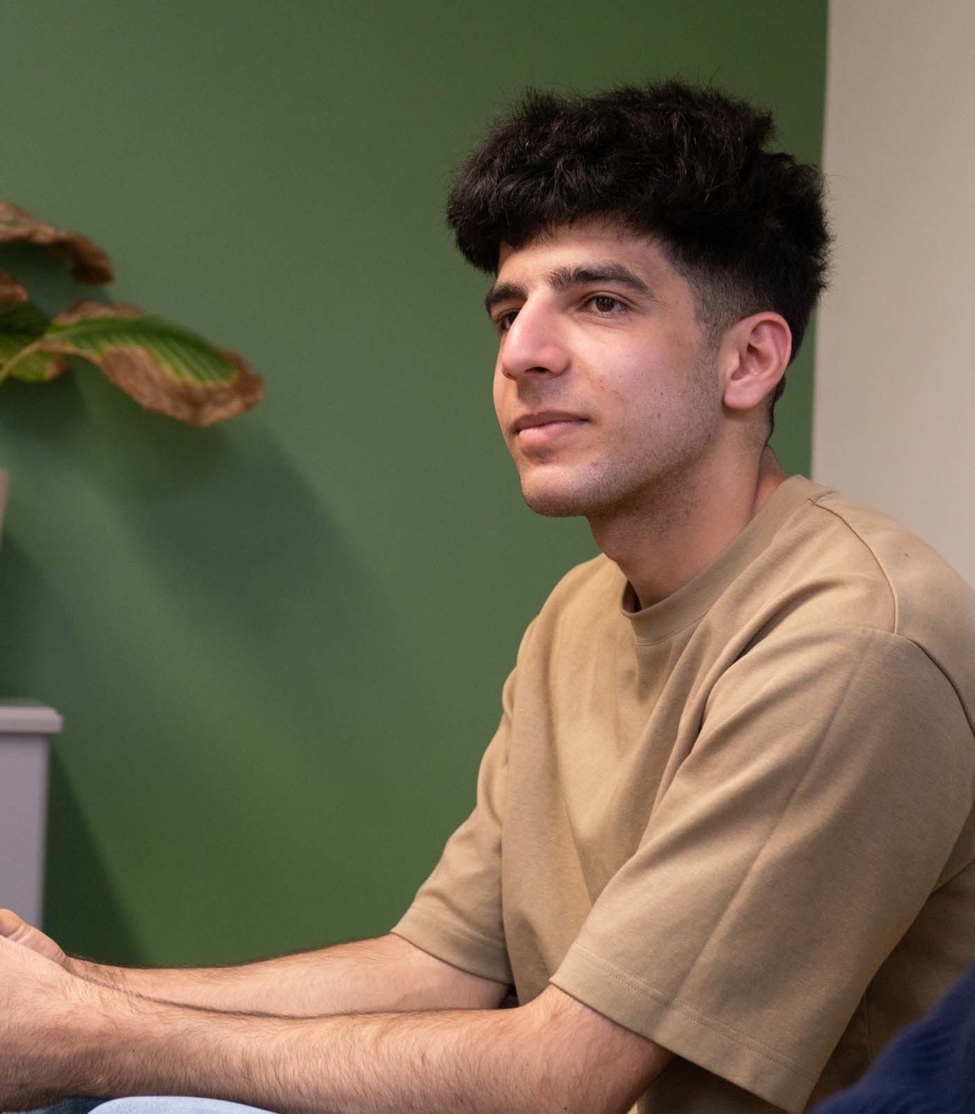 A young man with short, dark hair sits indoors against a green and beige wall. He is wearing a beige t-shirt and light blue jeans, looking ahead with a neutral expression. A plant is partially visible in the background.