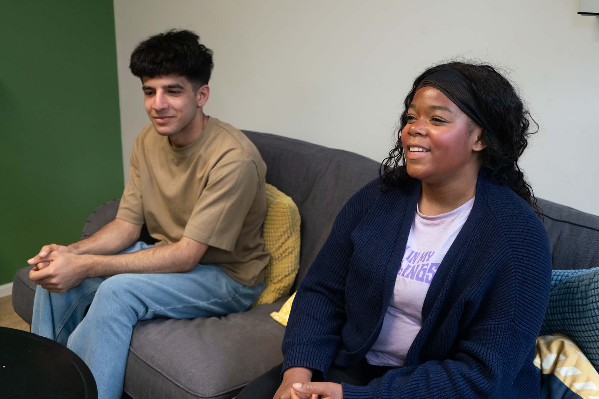Two people sit on a dark grey sofa, one wearing a tan shirt and jeans, the other in a navy cardigan and lavender shirt. They are facing forward and appear to be having a conversation. Yellow cushions are on the sofa.