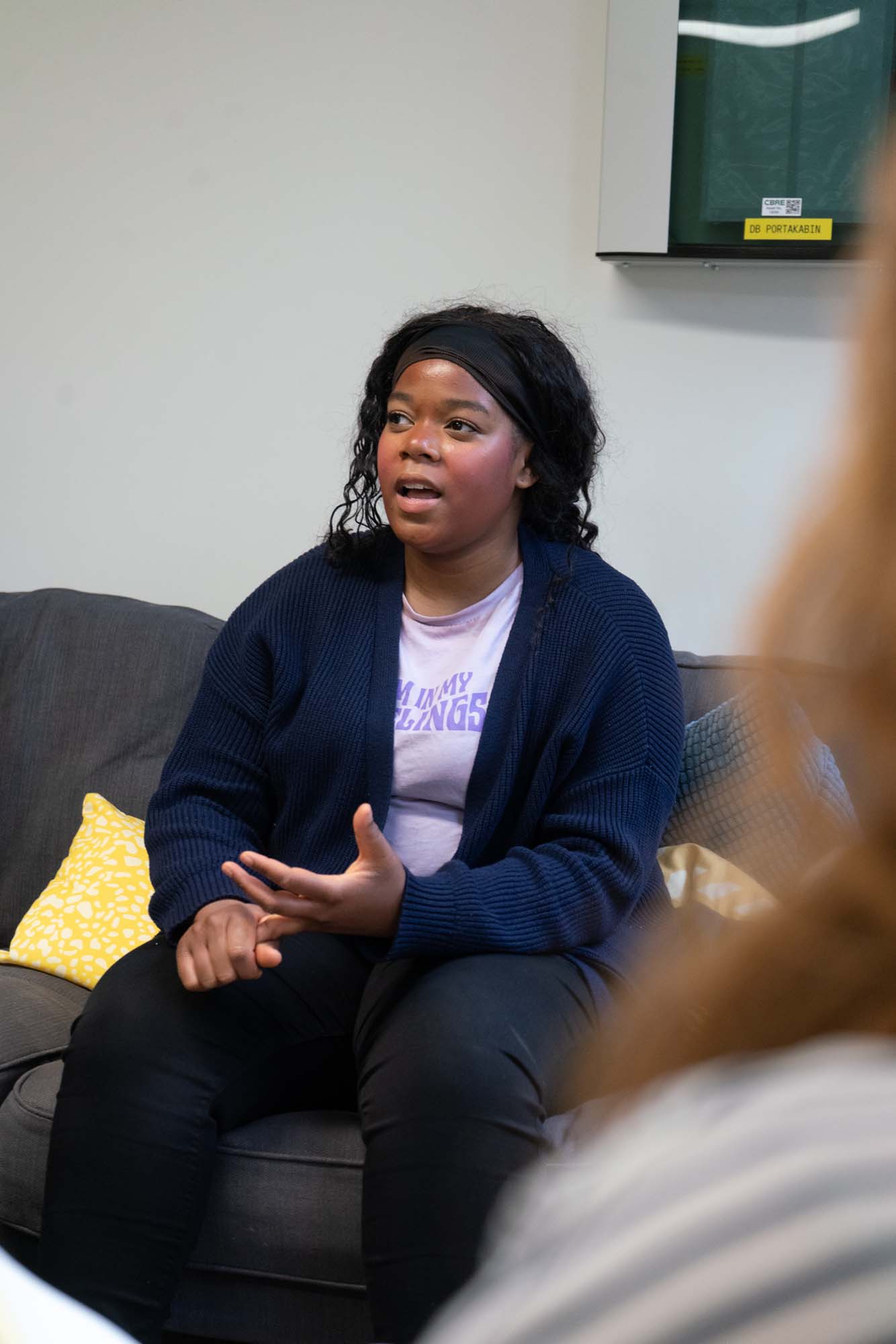 A person sits on a settee, gesturing with one hand while speaking. They wear a dark headband, navy jumper, and a light purple shirt. A yellow cushion and another seated person are partially visible.