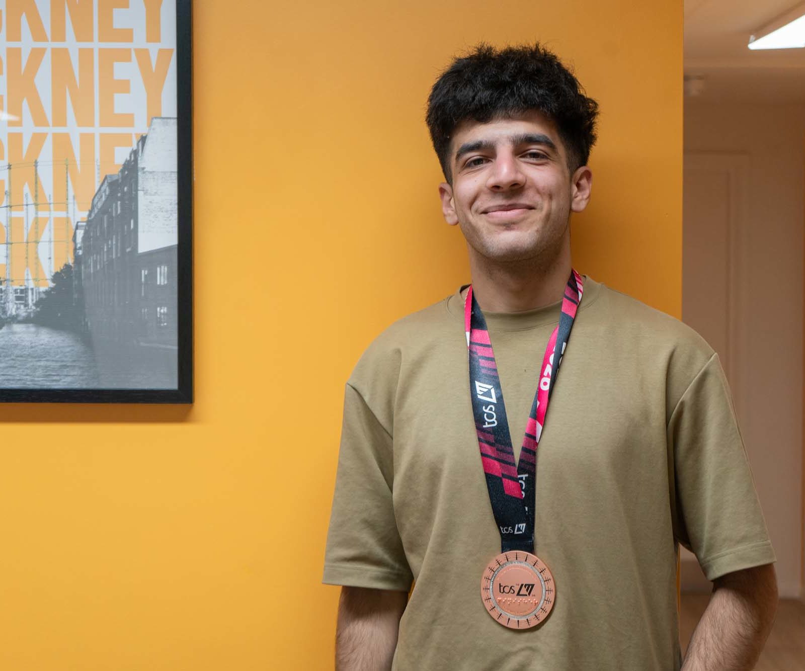 A young man with short dark hair stands indoors against an orange wall, wearing a tan t-shirt and a race medal around his neck. A framed HACKNEY poster is visible on the wall beside him.