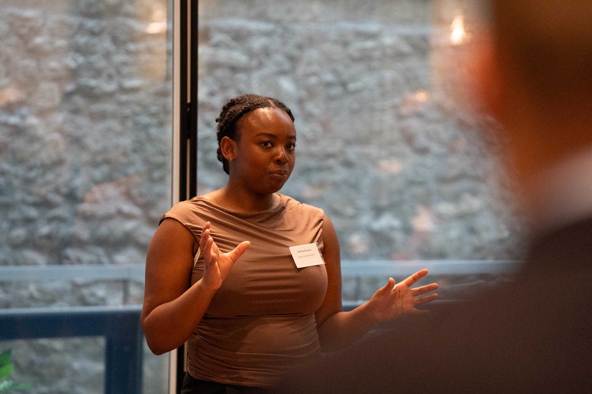 A woman wearing a brown top and name badge, gestures with her hands while speaking indoors. A blurred person is visible in the foreground.
