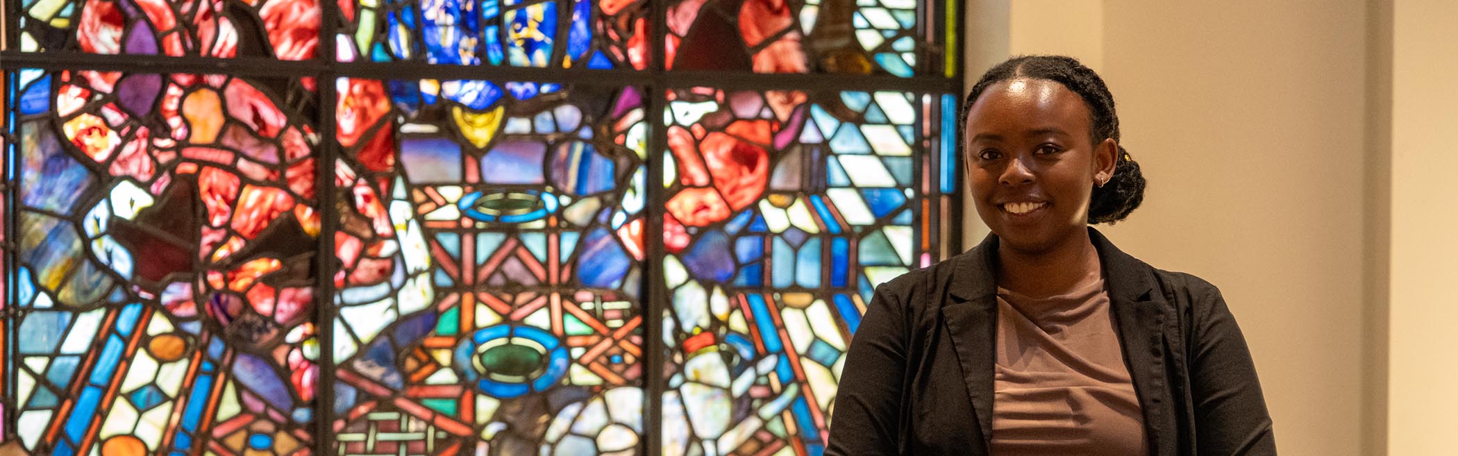 A woman with dark hair tied back, wearing a brown top and black jacket, stands indoors in front of a colourful stained glass window.