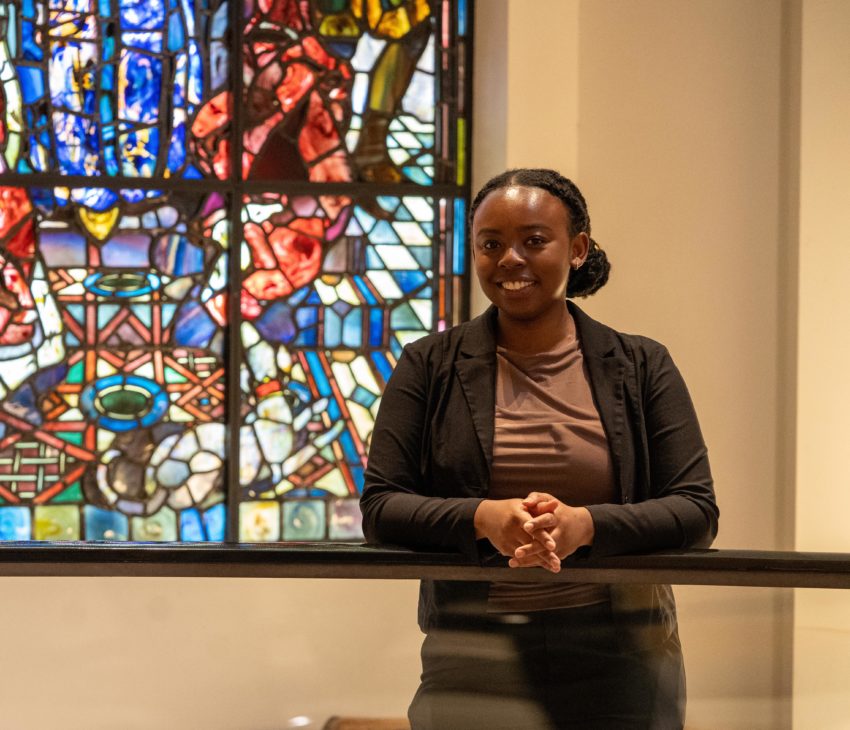 A woman stands indoors in front of a colourful stained glass window, resting her arms on a railing and smiling at the camera. She wears a dark blazer over a brown top.