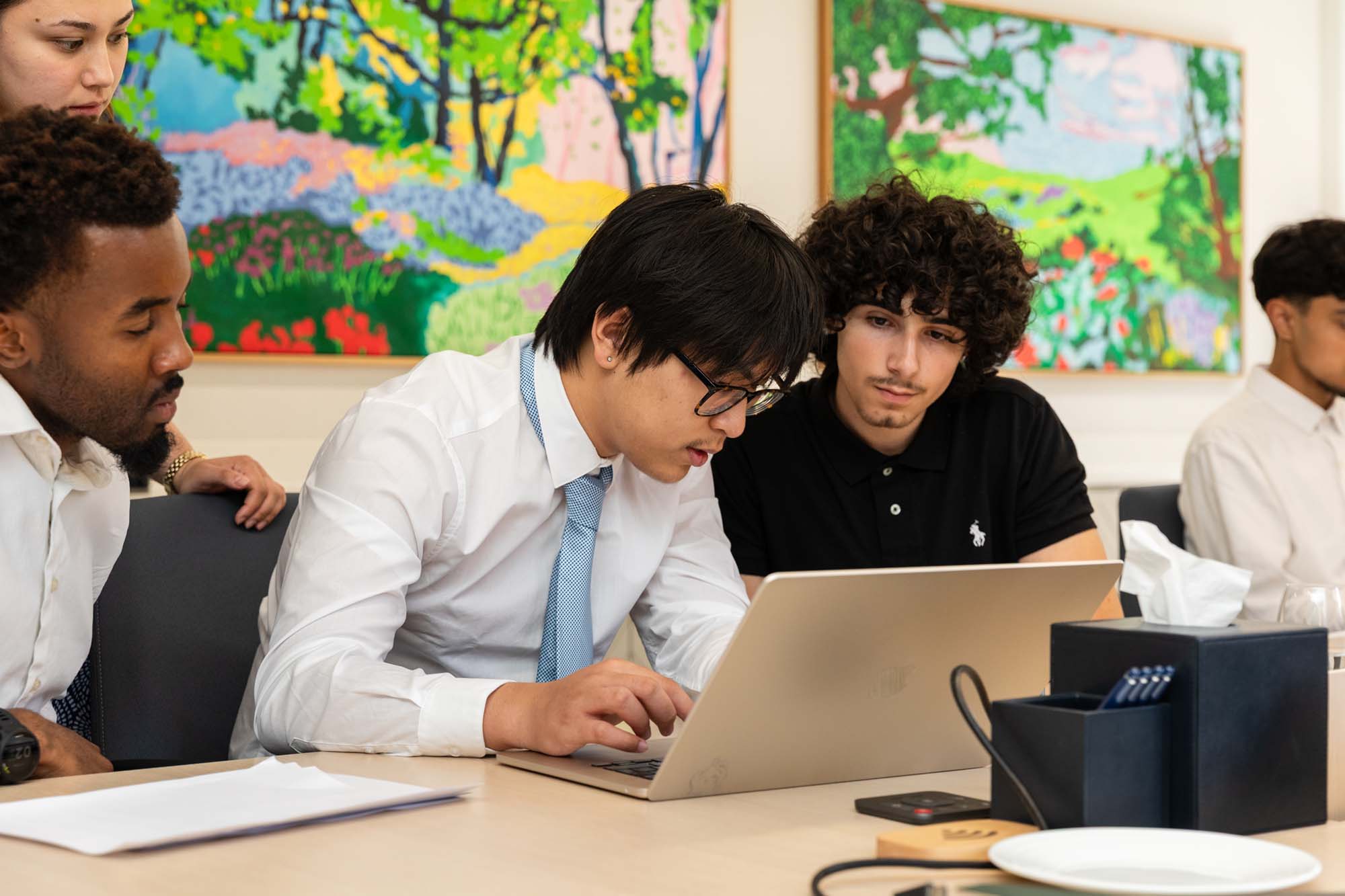 Four young men sit at a table. One types on a laptop while the others look on. Colourful artwork hangs on the wall behind them. Documents, a phone, and a tissue box are on the table.