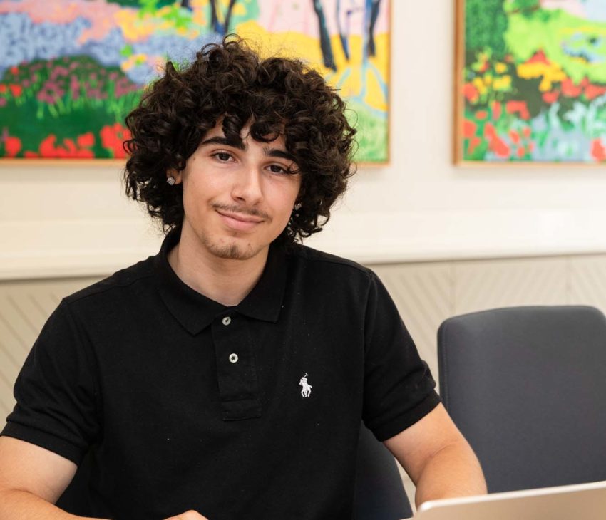 A young person with curly hair sits at a table with an open laptop, wearing a black polo shirt. Two colourful landscape paintings hang on the wall in the background.