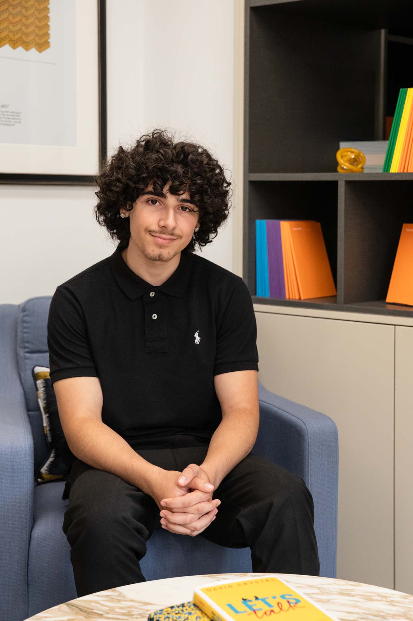 A person with curly hair wearing a black polo shirt sits on a blue chair with hands clasped. Colourful books and decor are arranged on shelves behind them. A book and part of a table are visible in the foreground.
